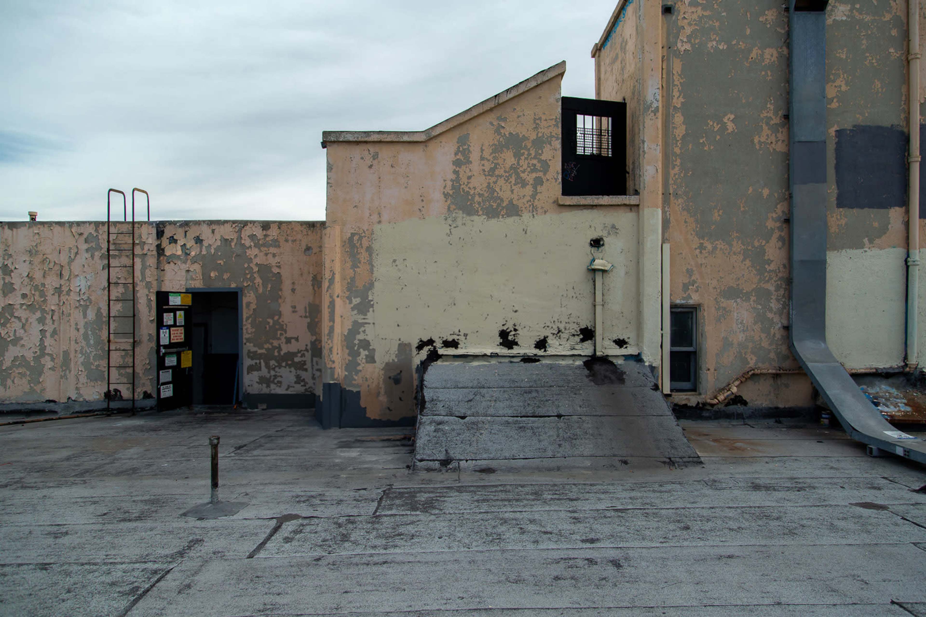 The image shows a rooftop with peeling paint, a metal ladder, an opening leading into a building, and a sloped structure next to a drainage pipe.