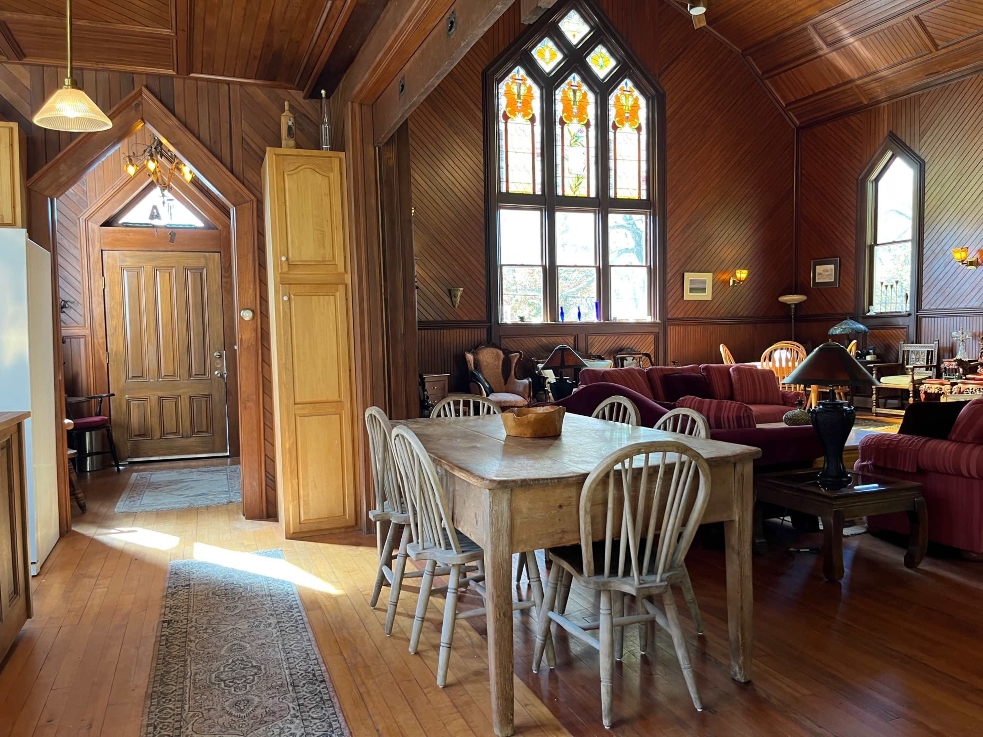 The interior of a wooden-paneled room features a dining table surrounded by chairs, a large entrance with a decorative door, and stained glass windows illuminating the space.