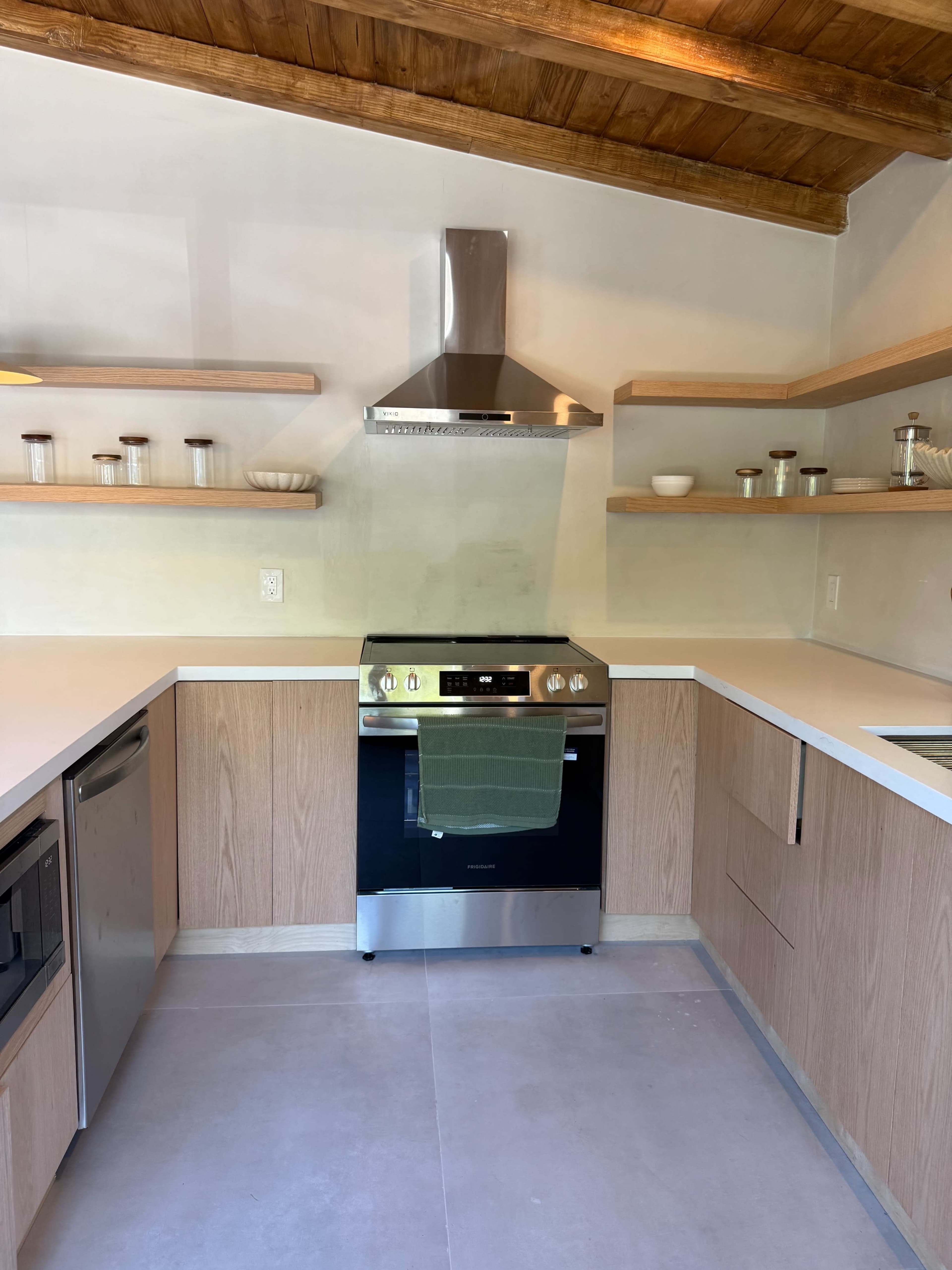The image shows a modern kitchen with wooden shelves, a stainless steel range hood, and an oven with a green towel hanging from the handle.