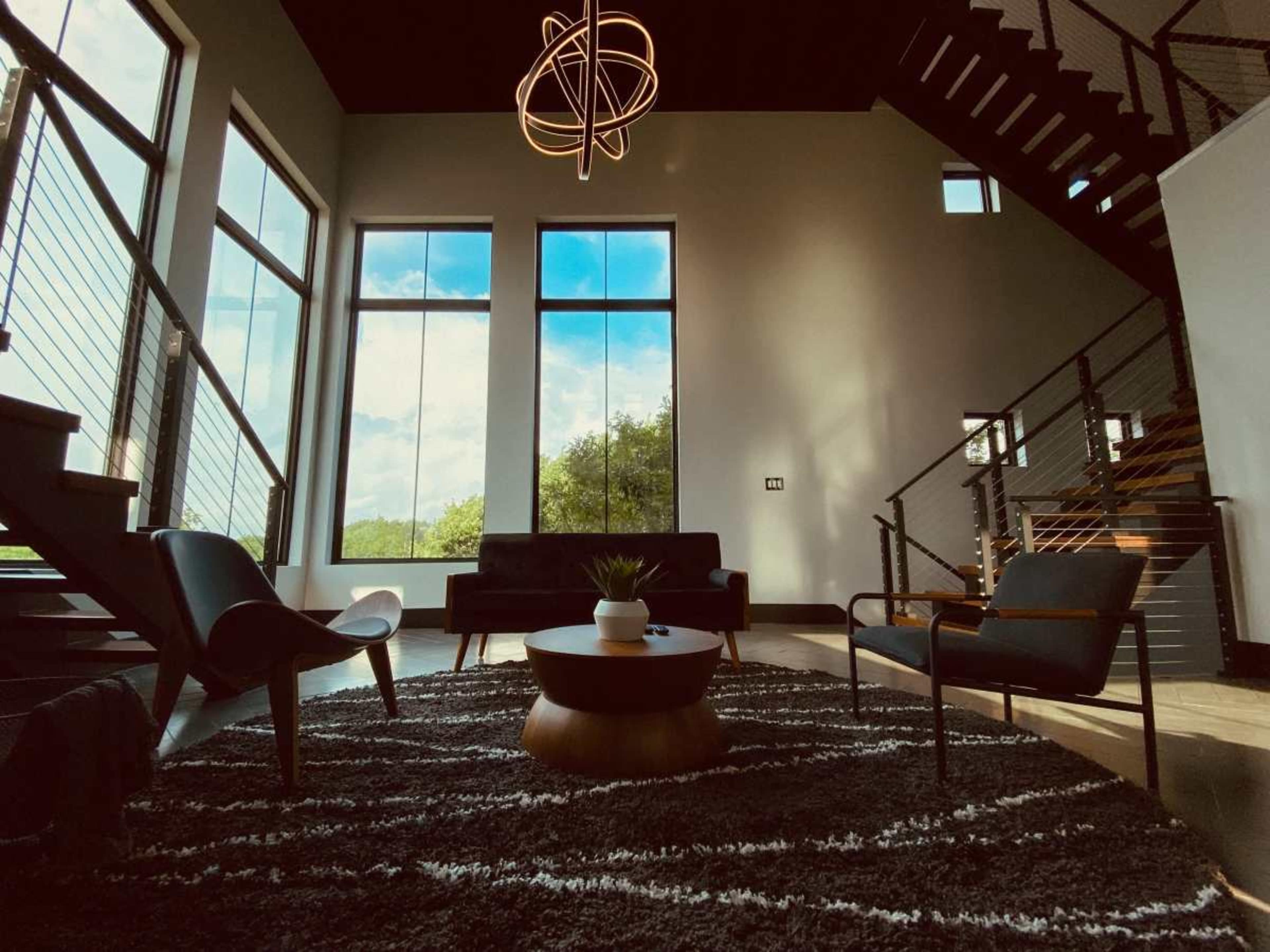 A spacious living area features large windows, modern furniture, and a staircase leading to an upper level.