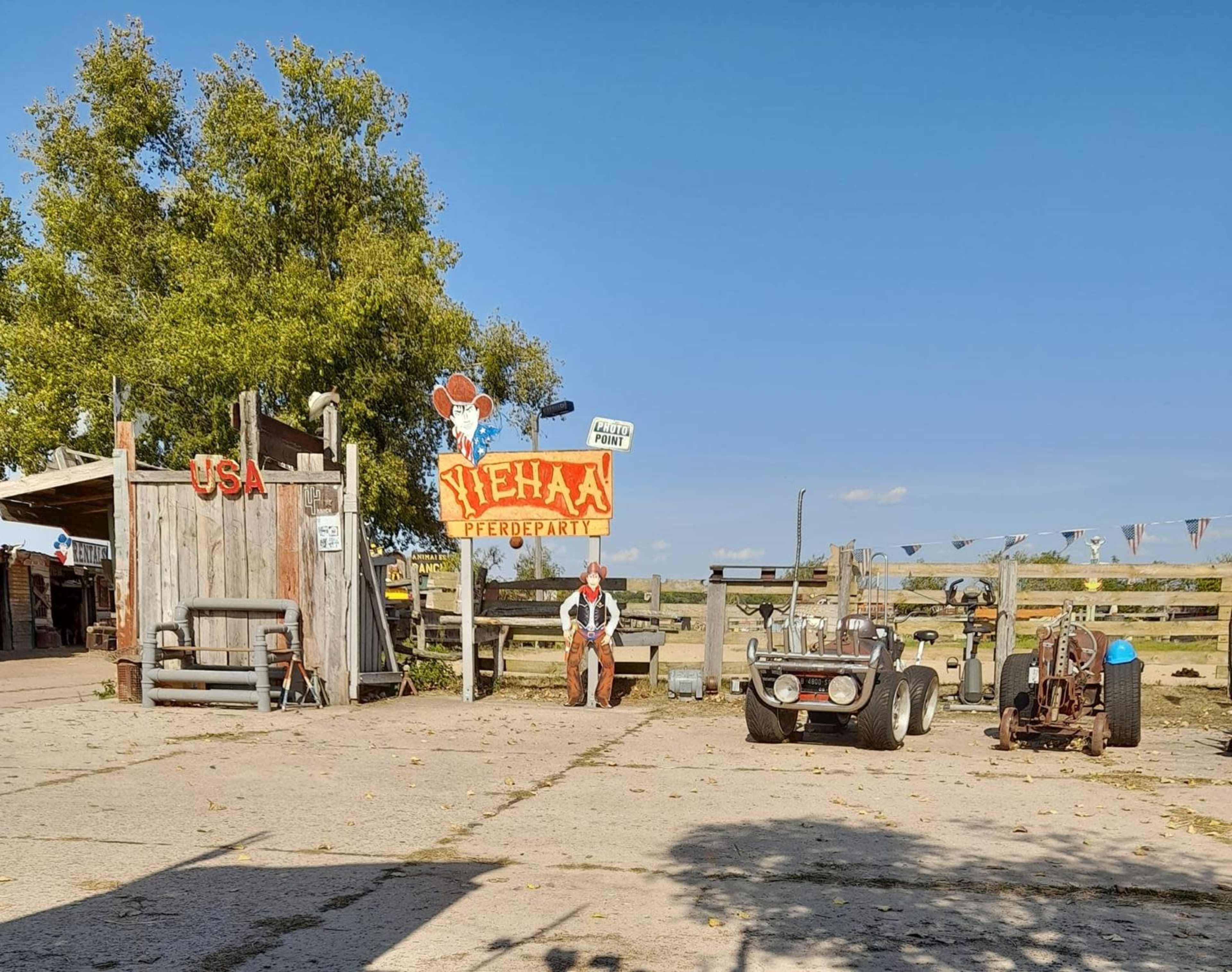 The image shows a rustic Western-themed setting with a vibrant sign reading "Vieha" and vintage cars parked in front of a wooden structure.