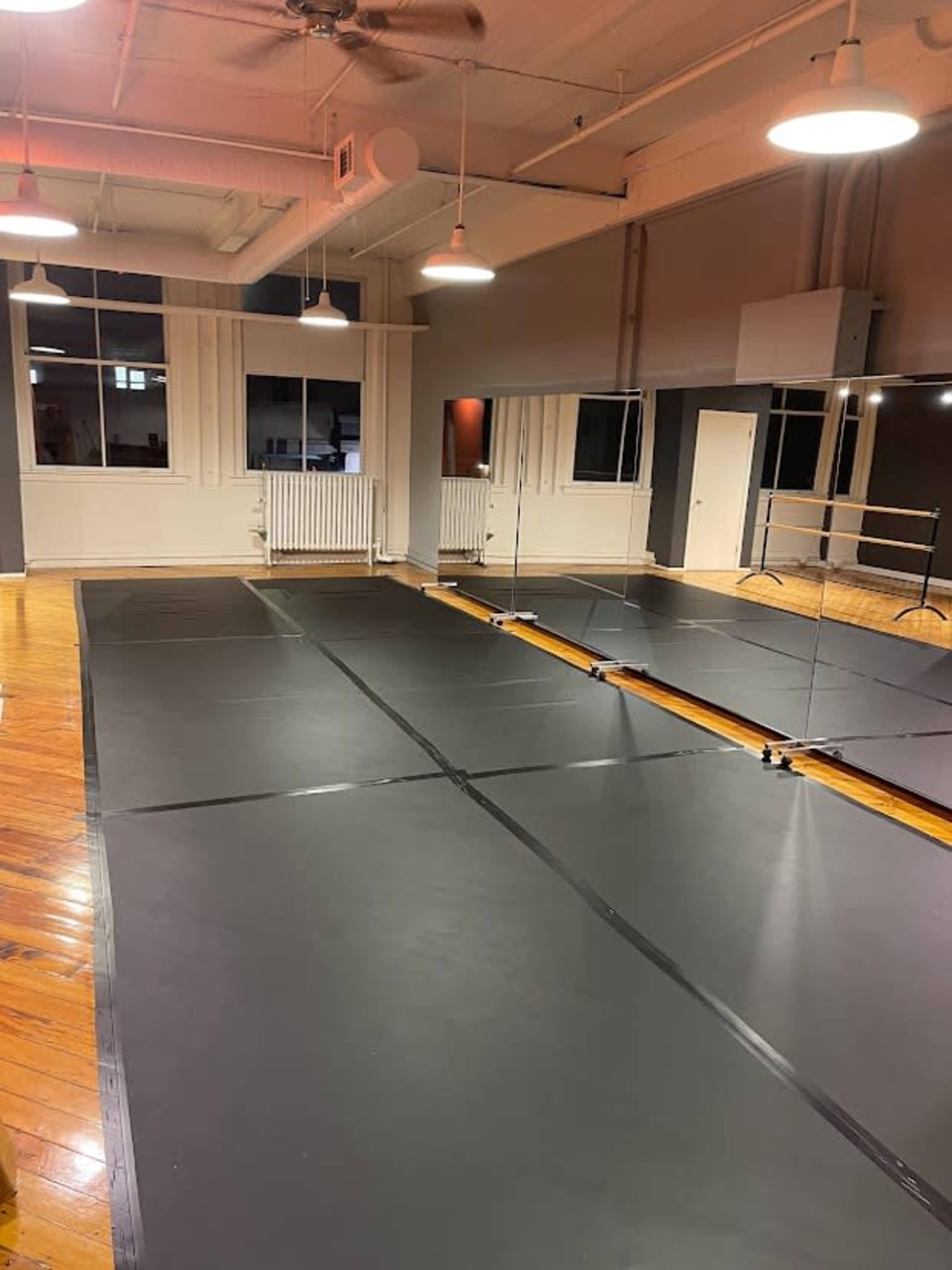 A dance studio featuring a black rubber floor, large mirrors on one wall, and overhead lighting.