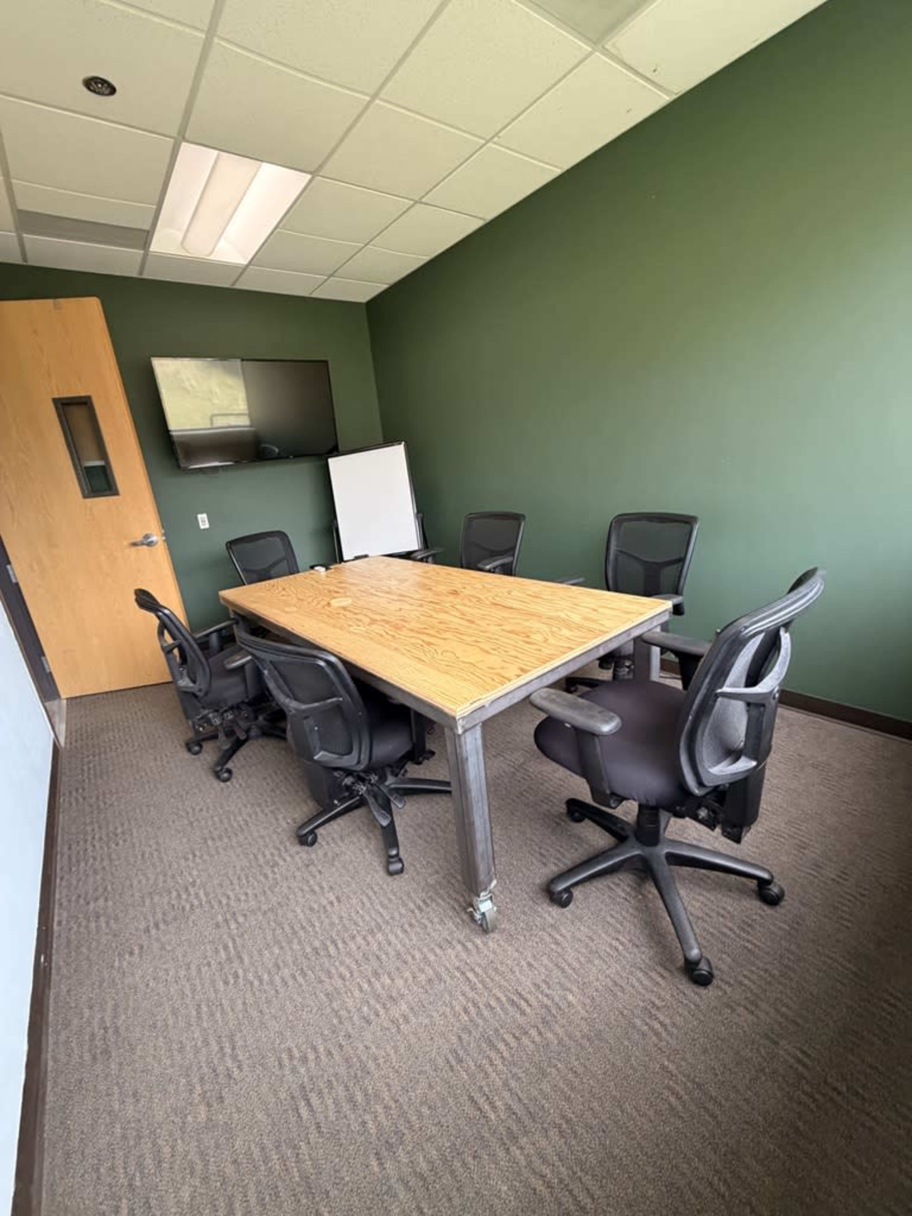 A conference room features a large wooden table surrounded by six black office chairs, with a television mounted on the wall and a whiteboard in the corner.
