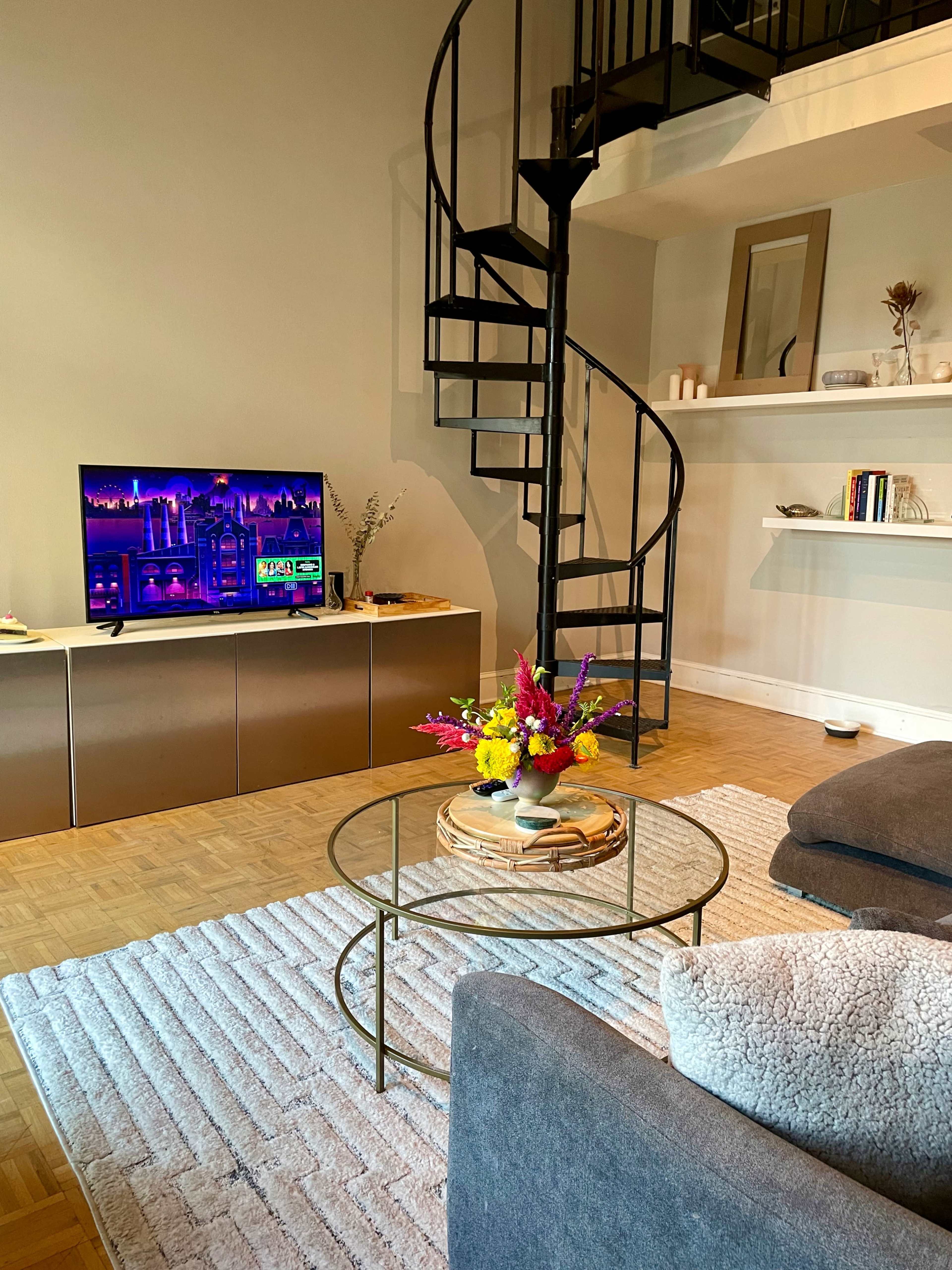 A living room features a spiral staircase, a television on a cabinet, a coffee table with a floral arrangement, and a gray sectional sofa on a textured rug.