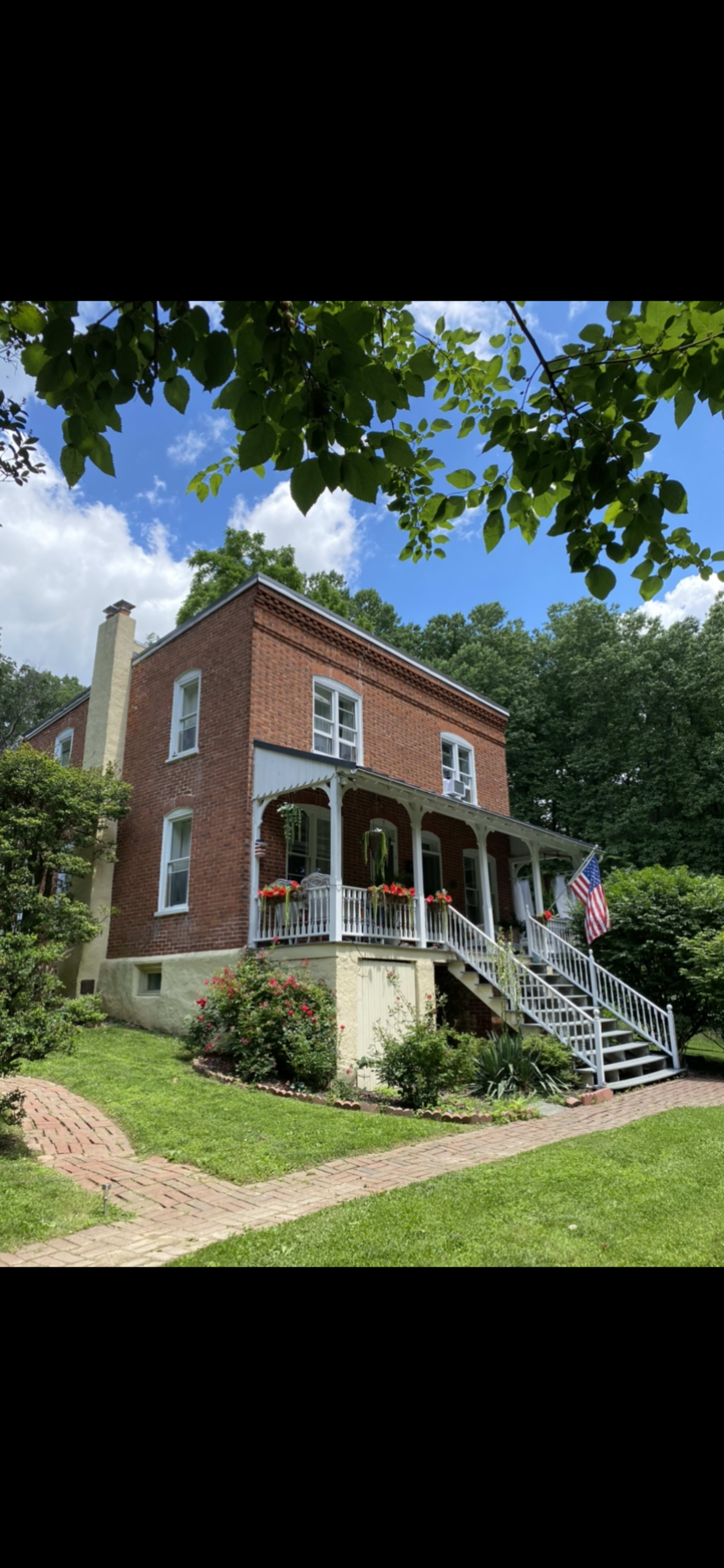 A two-story brick house with a white railing porch is surrounded by green grass and flowering plants.