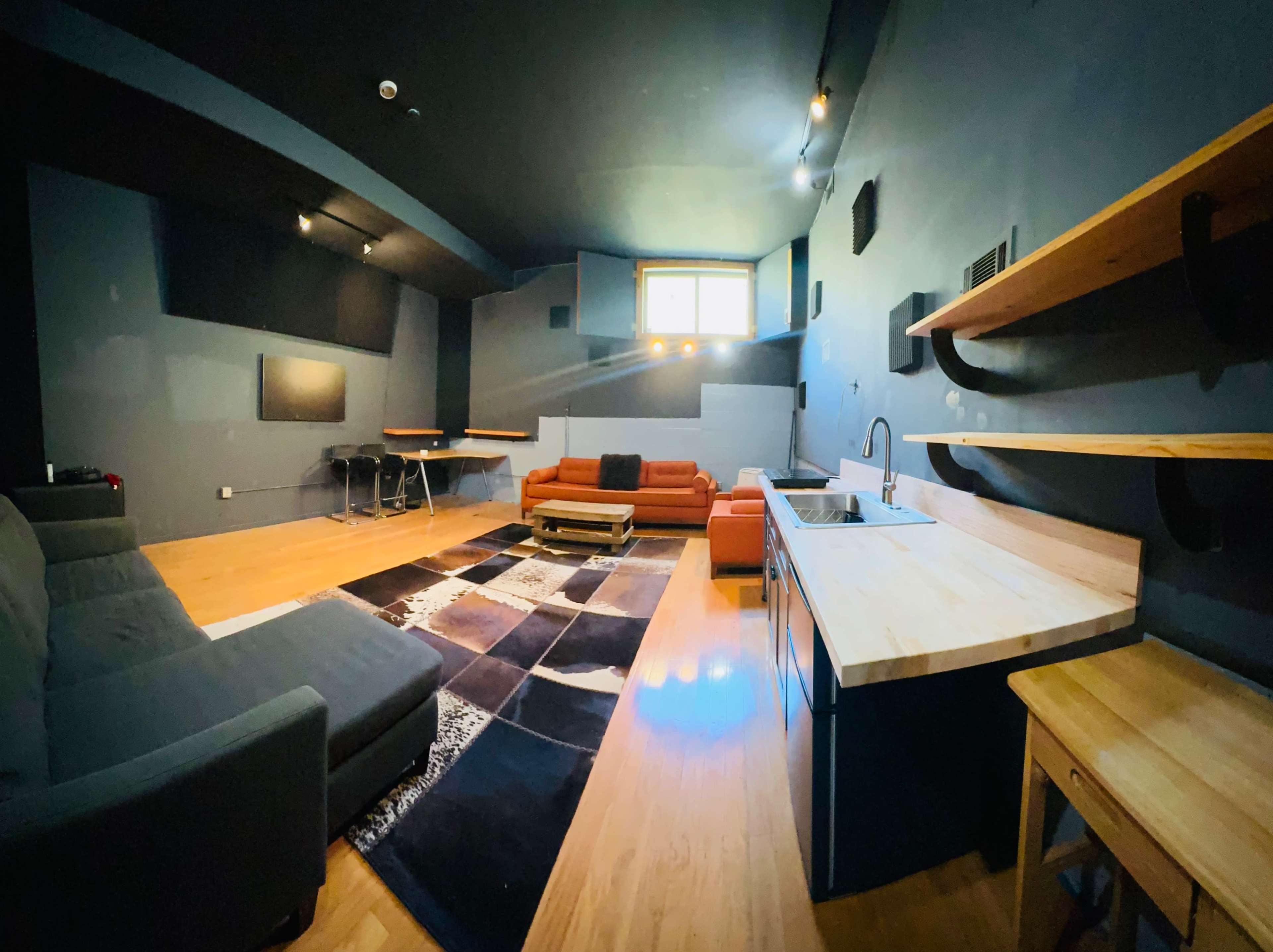 The image shows a modern interior room furnished with orange sofas, a kitchenette, a large rug, and a black dining table.