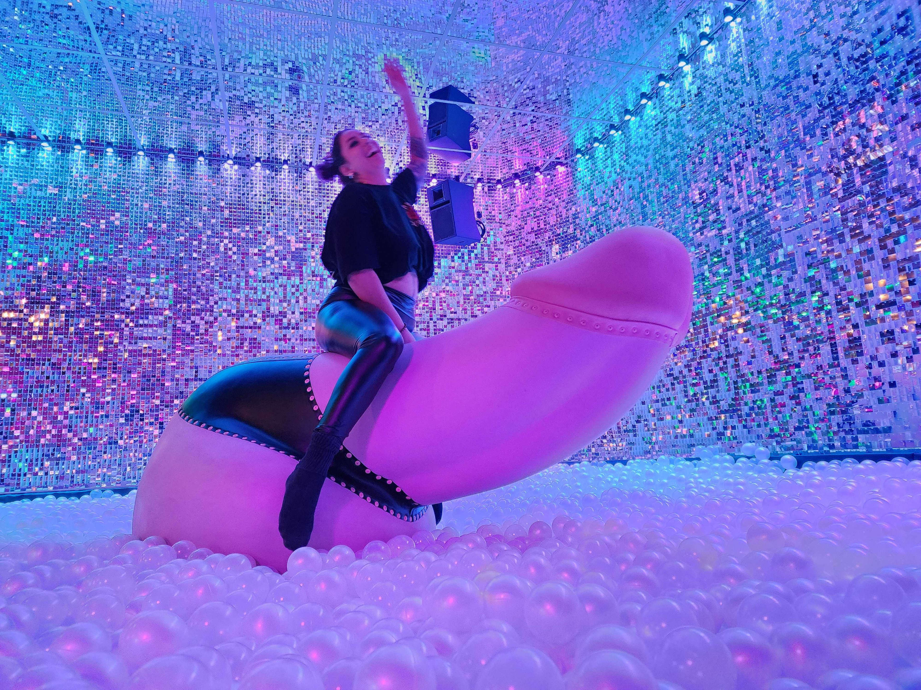 A person is sitting on a large, pink sculpture shaped like a phallus in a colorful, mirrored room filled with pink bubbles.