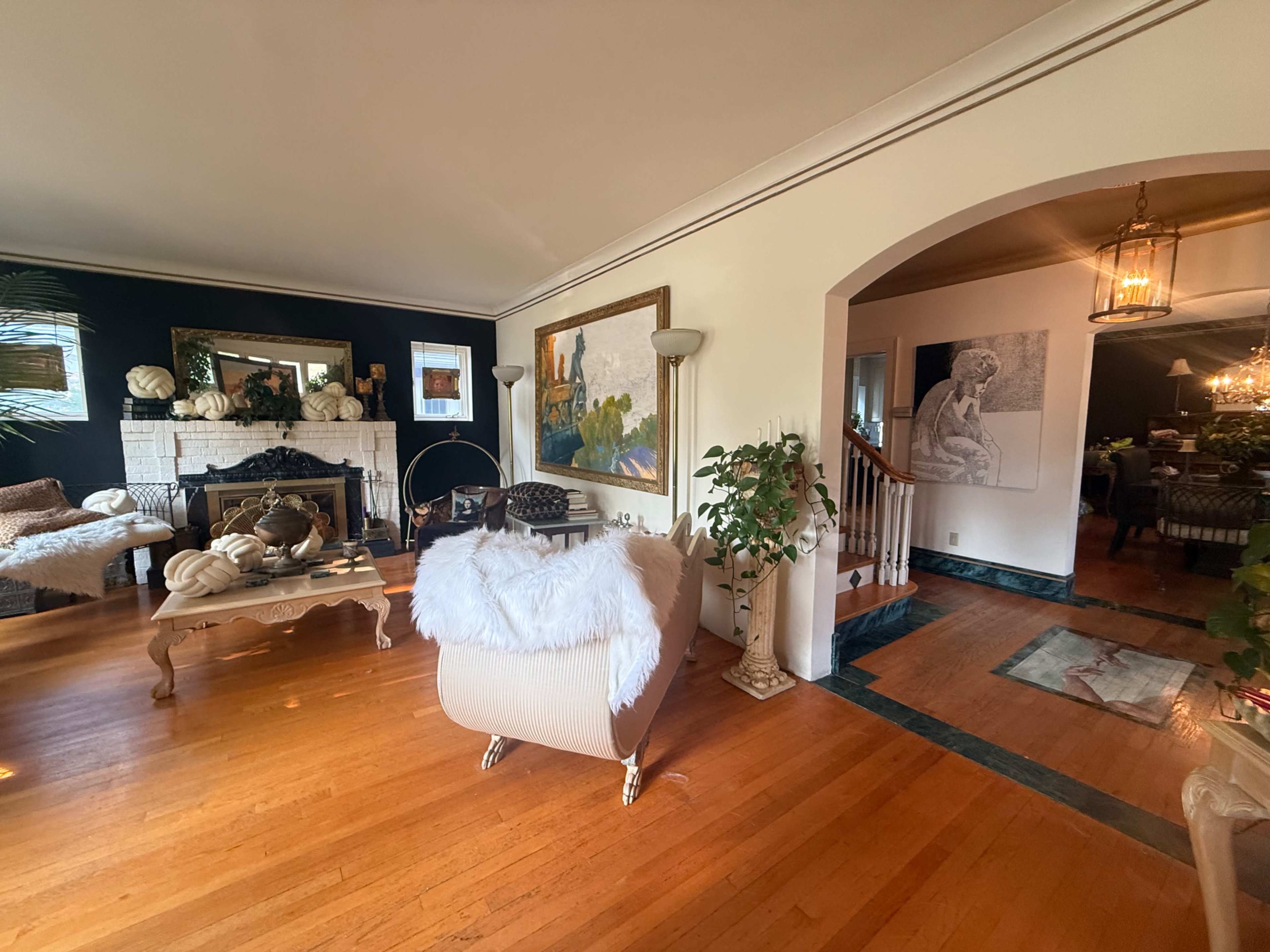 The image depicts a spacious living room featuring hardwood floors, a fireplace, decorative artwork, and a staircase leading to an adjacent dining area.