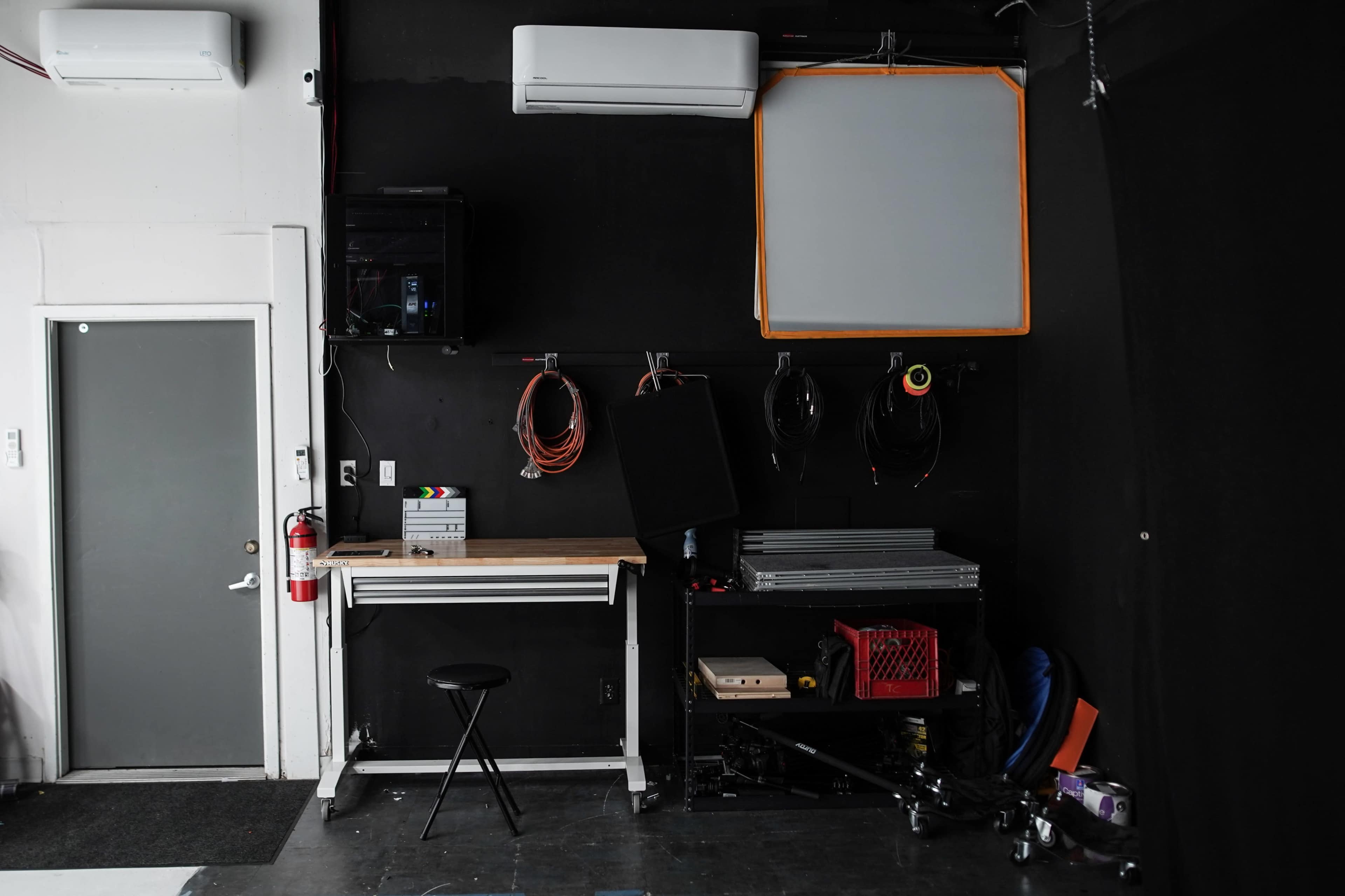 A minimalist studio space featuring a worktable, stool, and various cables hanging on a black wall beside a door and equipment storage.