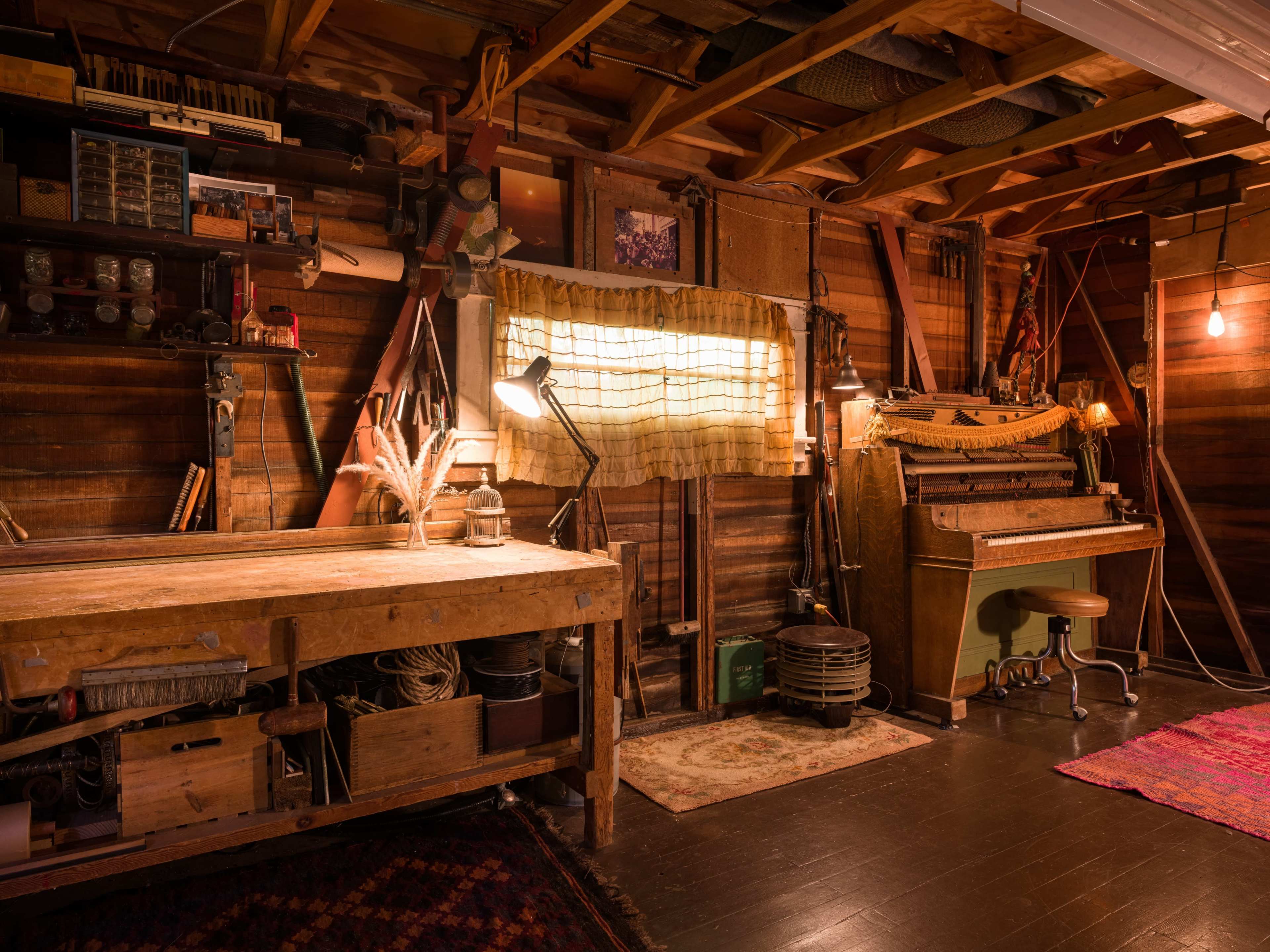The image depicts a rustic workshop with a wooden workbench, an antique piano, and soft lighting illuminating the space.