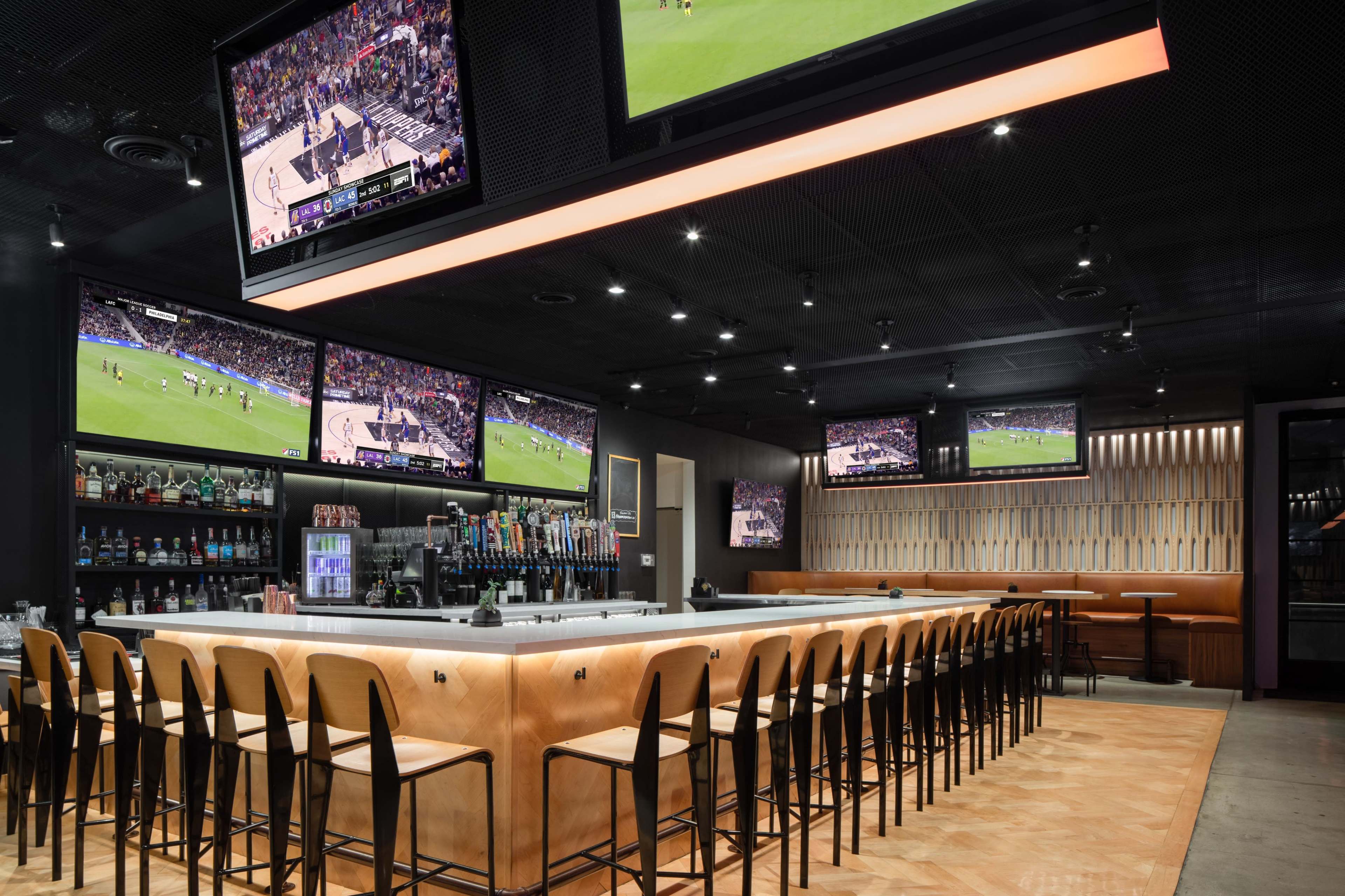 The image shows a modern bar area with a long counter and multiple large televisions displaying sports games.