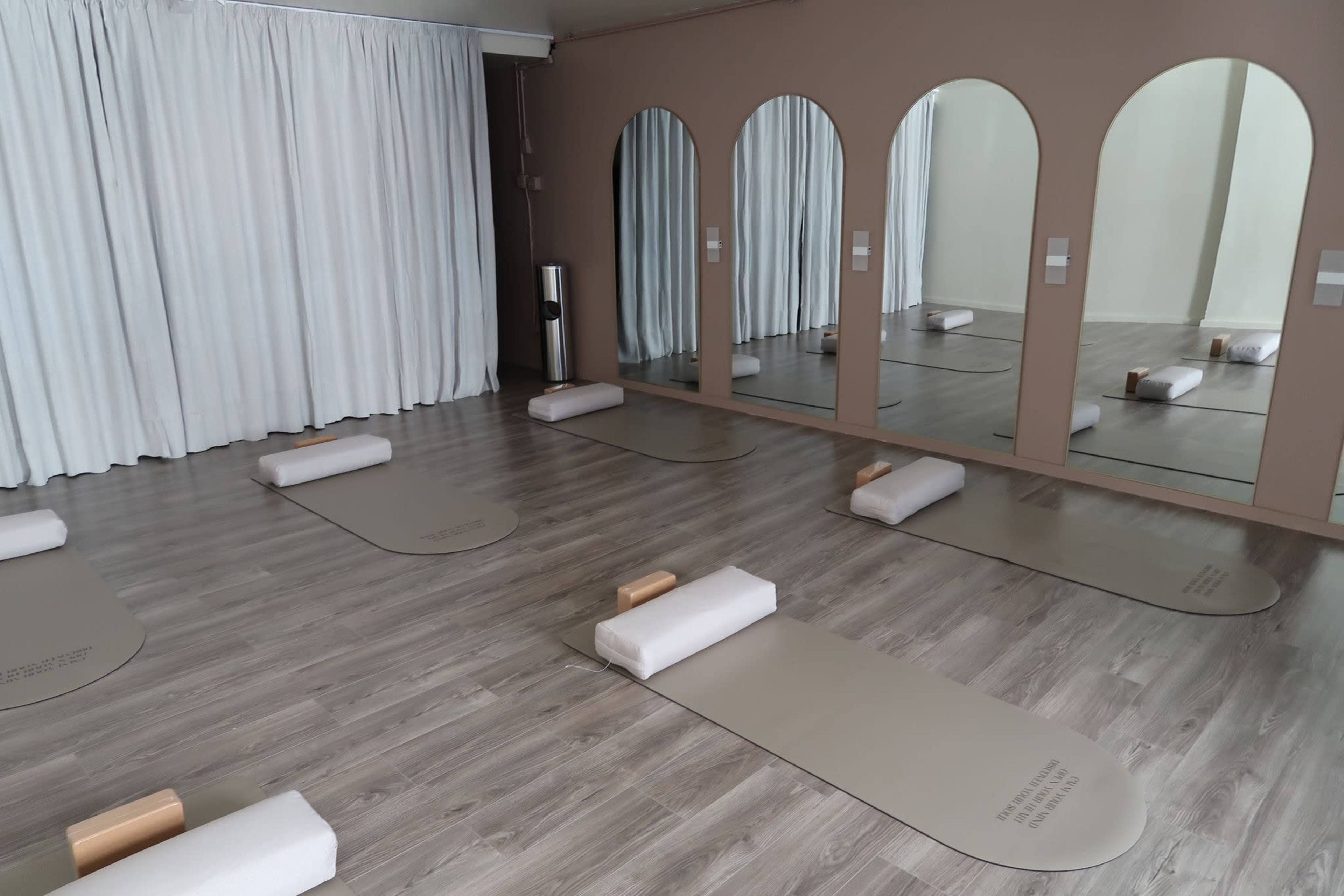 The image shows a minimalistic studio space with several exercise mats arranged on a wooden floor, mirrors on the wall, and rolled towels placed on each mat.