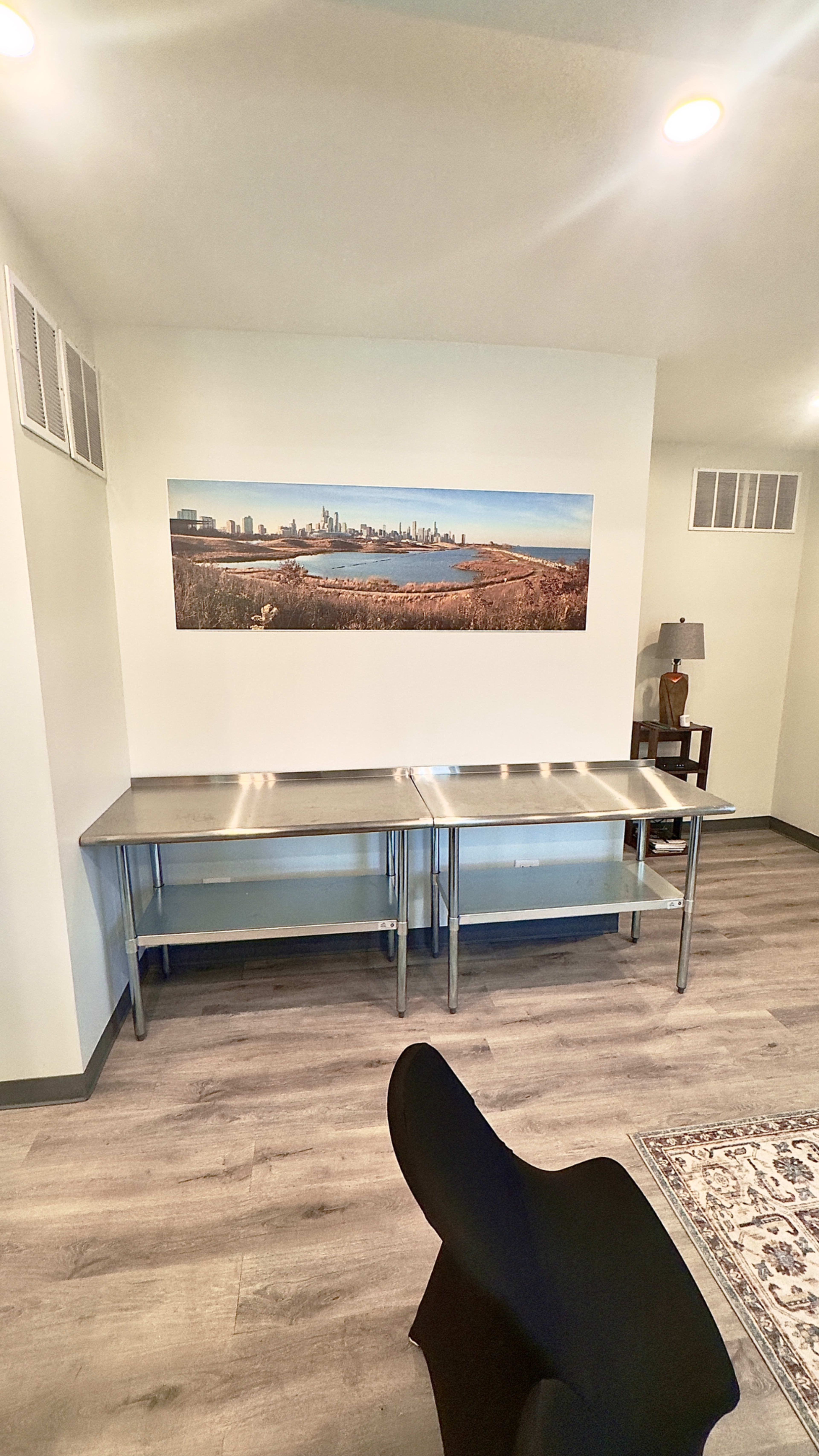 The image shows a room with a long stainless steel table against a wall featuring a panoramic photograph of a city skyline, alongside a table lamp and a small chair.