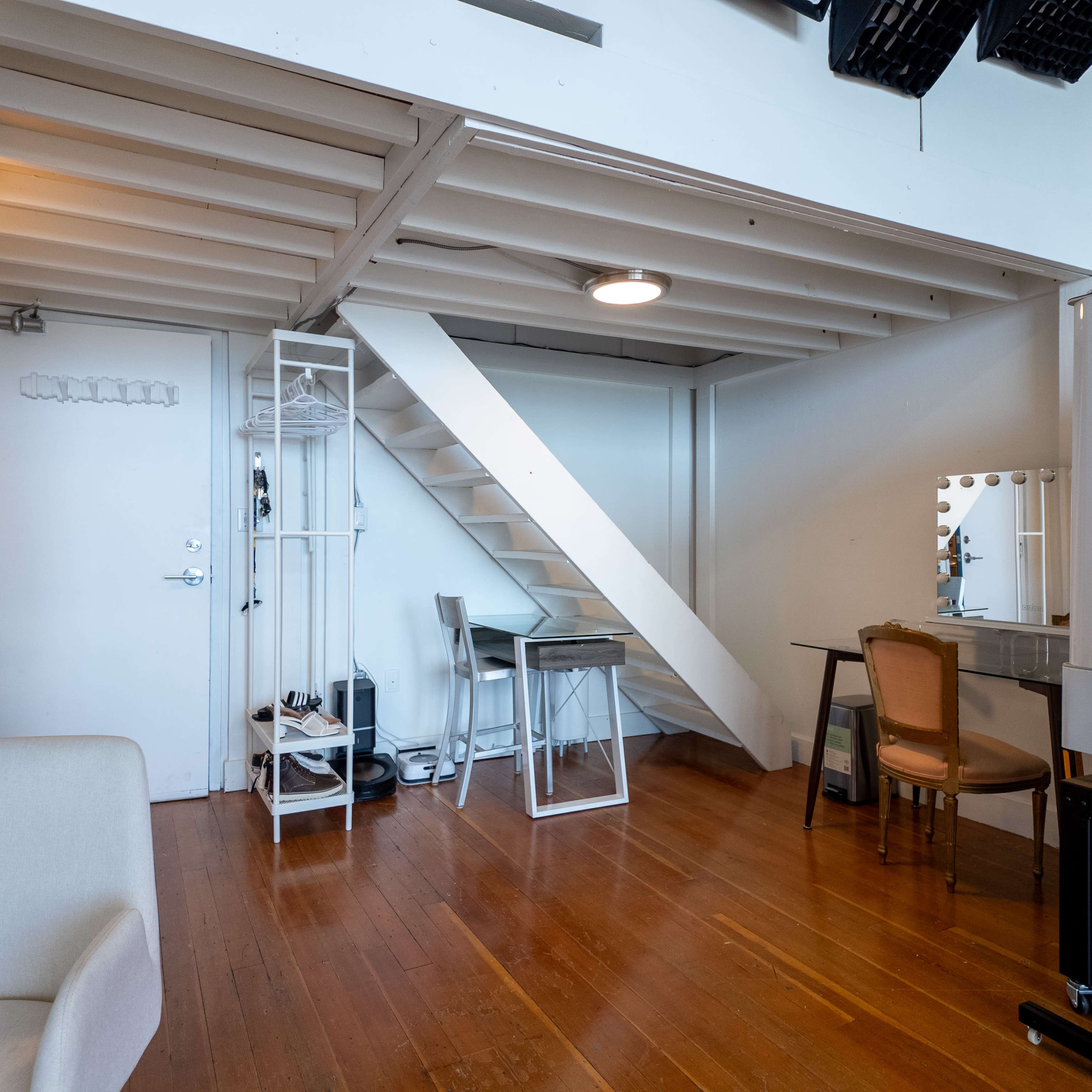 A bright, minimalistic room with a staircase leading to an upper level, a small workspace, and a dressing table along the walls.