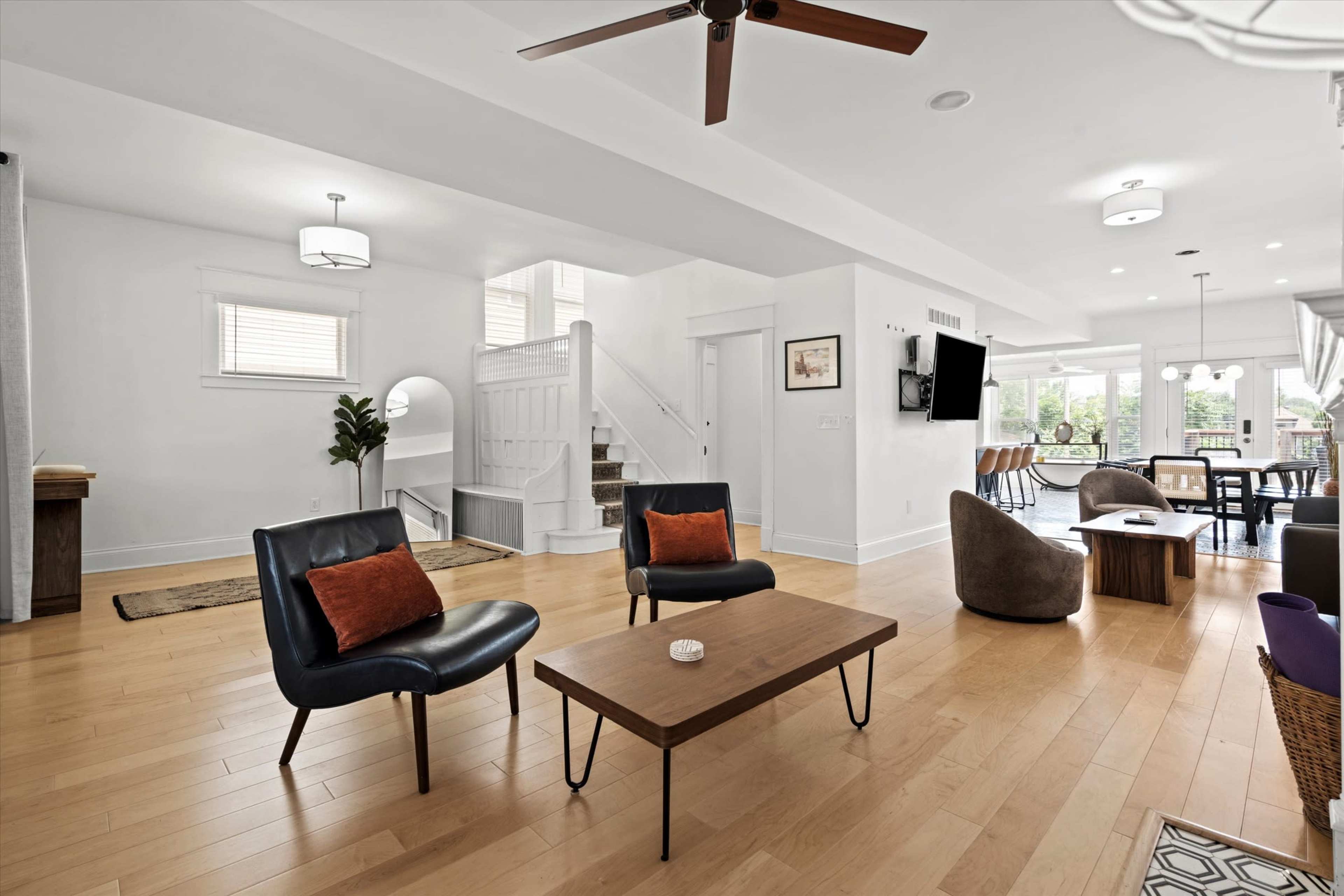 The image shows a modern, open-concept living room with seating arrangements that include two chairs with cushions, a coffee table, and a view of the dining area and staircase.