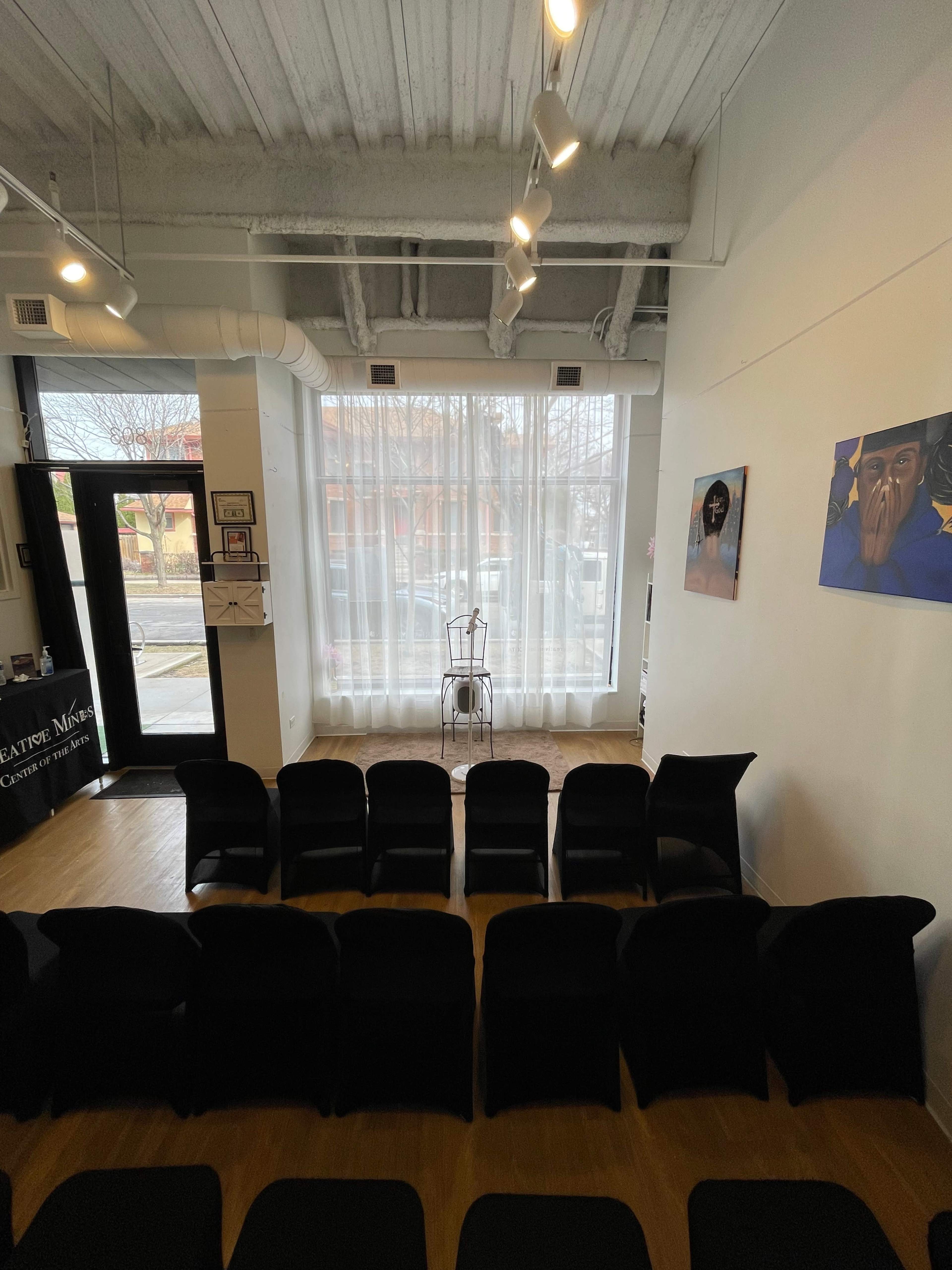 The image shows a minimalist waiting room with black chairs arranged in rows facing a bright window and a small stage with a stool.