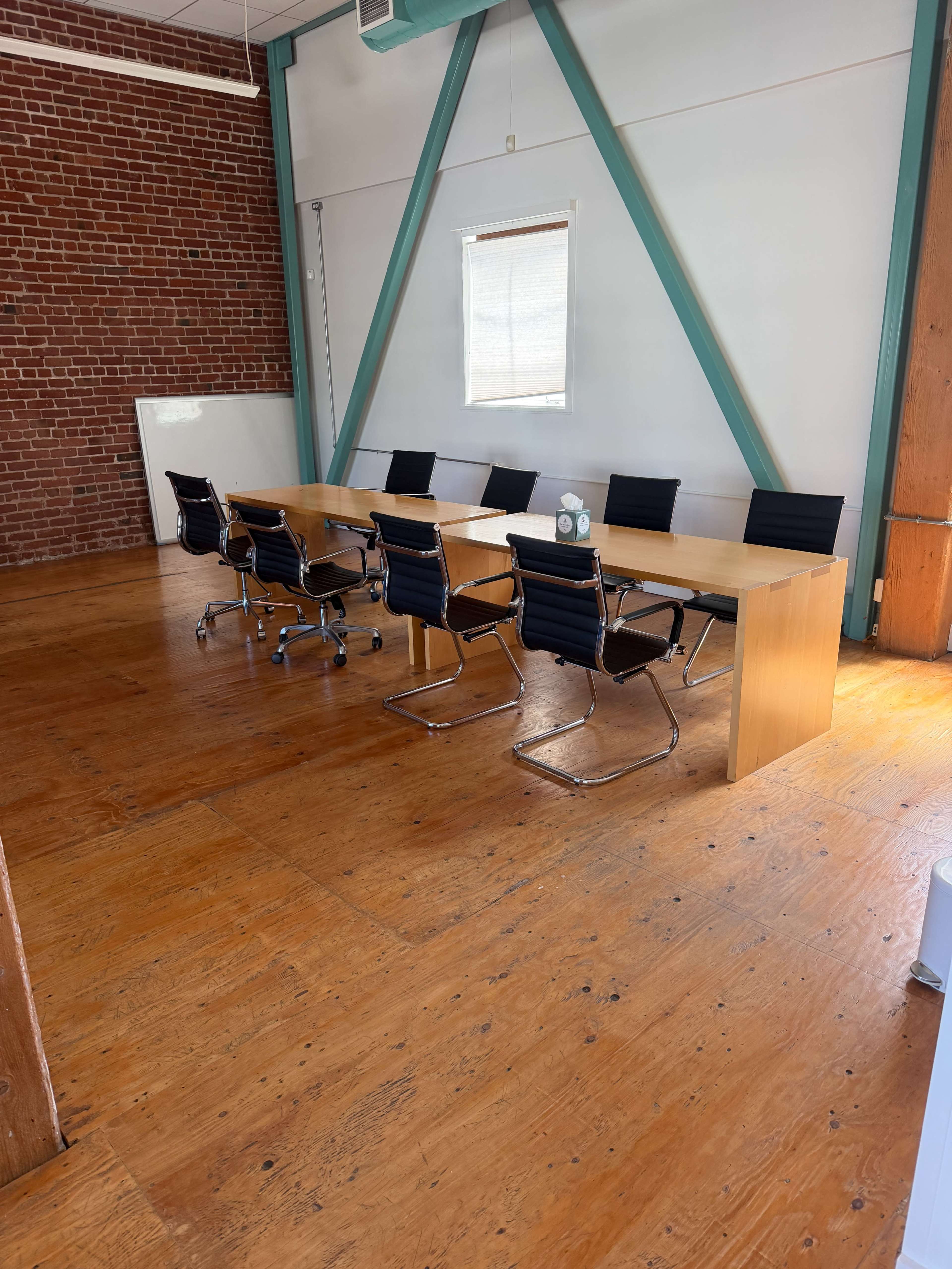 A long wooden conference table with eight black chairs is set up in a spacious room featuring exposed brick walls and wooden flooring.