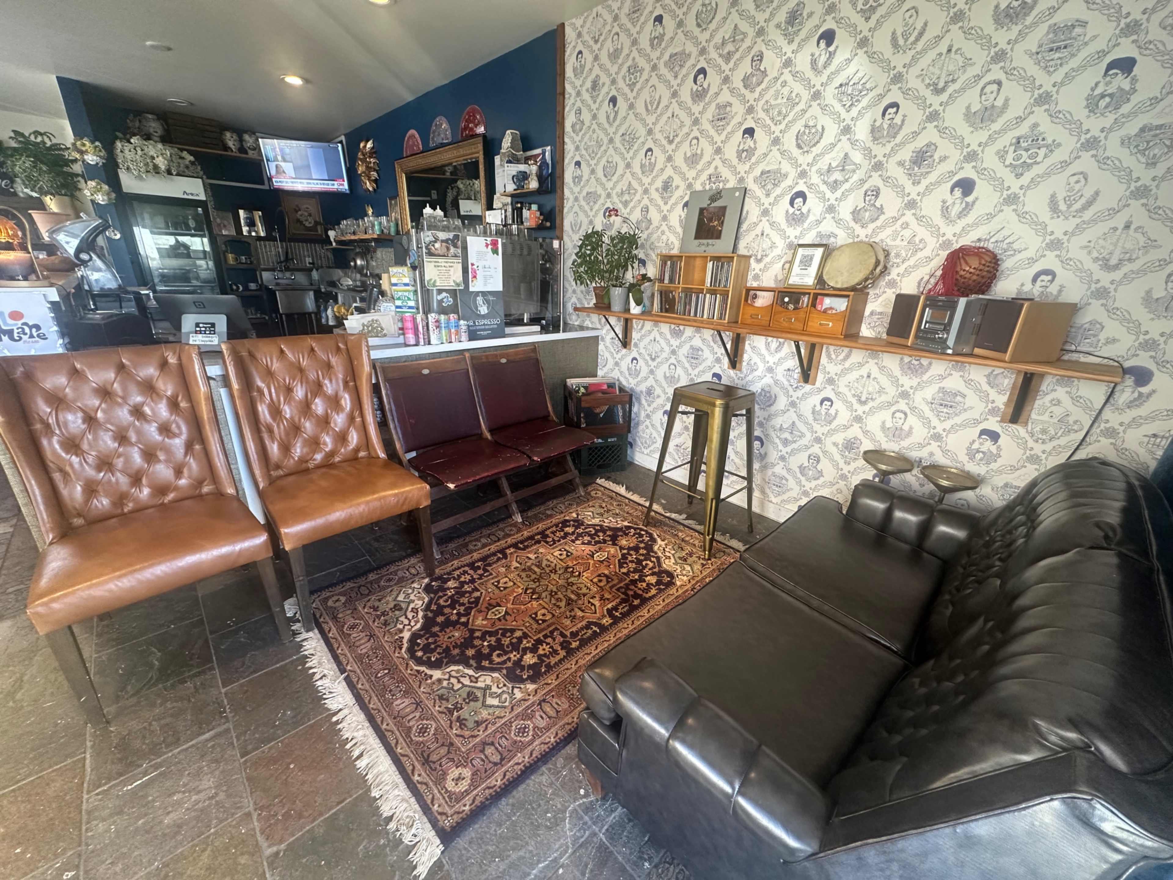 The image shows a cozy café interior featuring a mix of brown leather chairs, a dark couch, an area rug, and a wall decorated with patterned wallpaper and shelves displaying various items.
