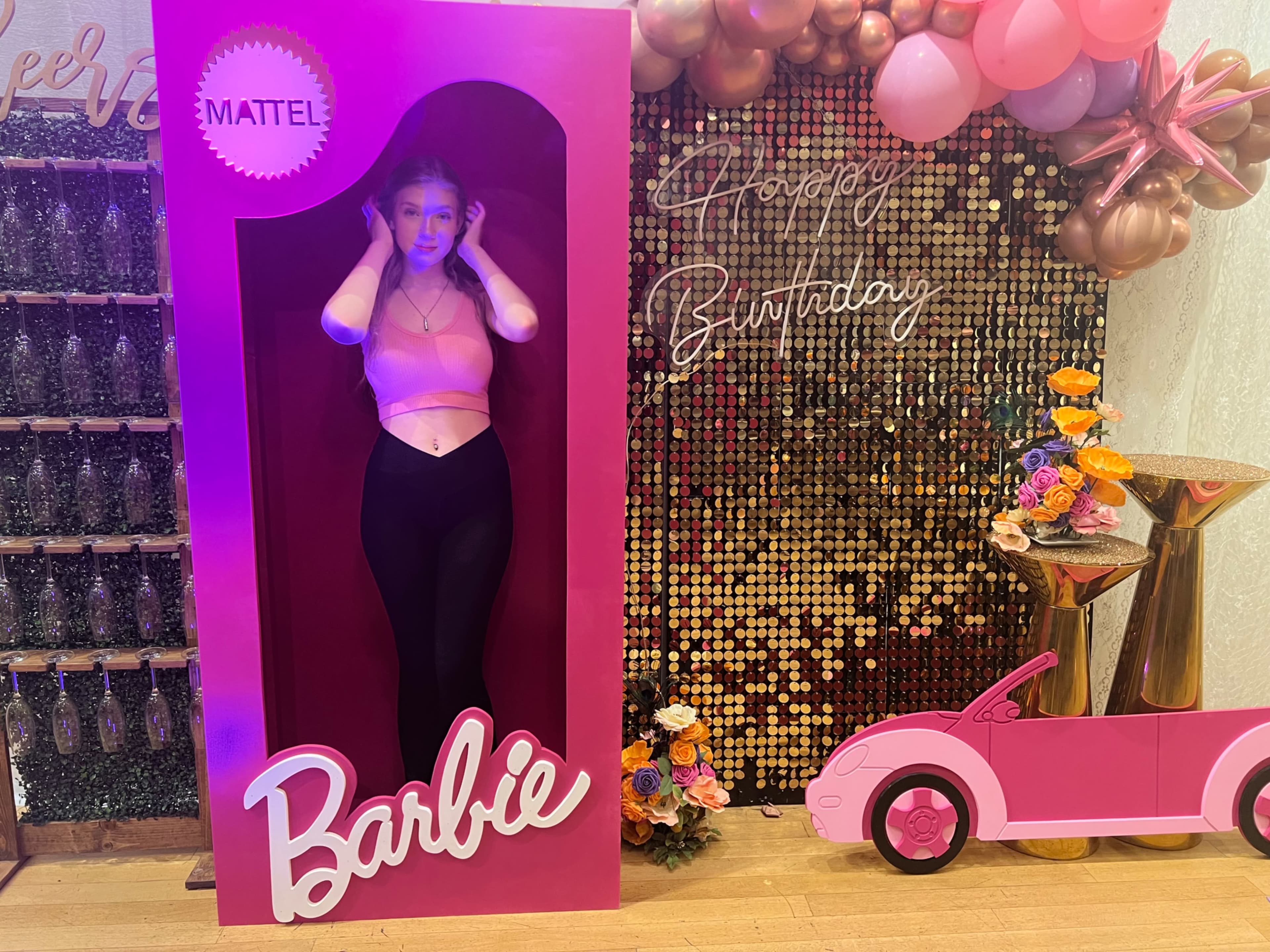 A person stands in a Barbie-themed display with a pink frame, surrounded by festive decorations for a birthday celebration.