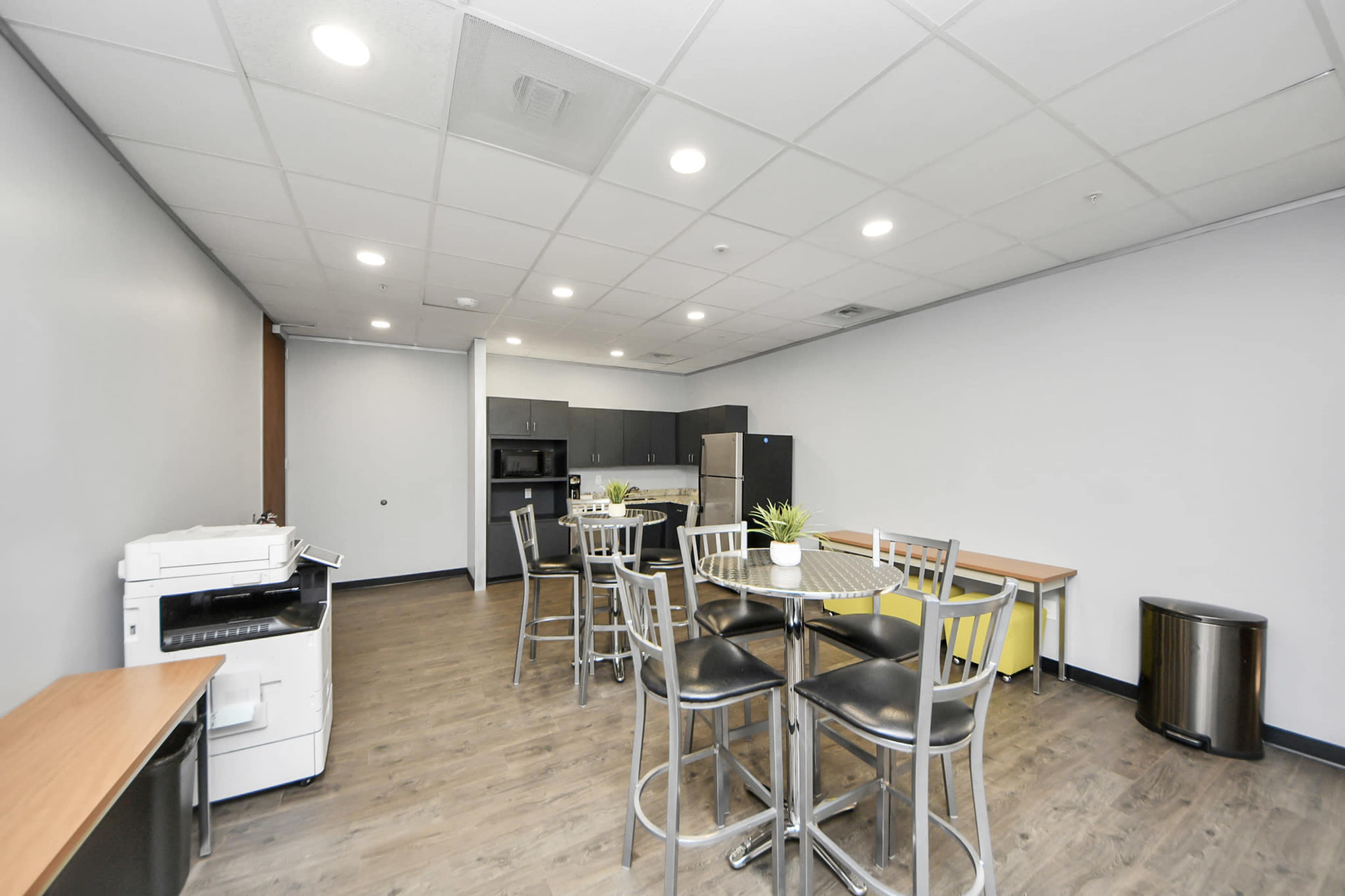 The image shows a break room with modern furniture, including a tall table with chairs, a kitchen area with appliances, and a printer.
