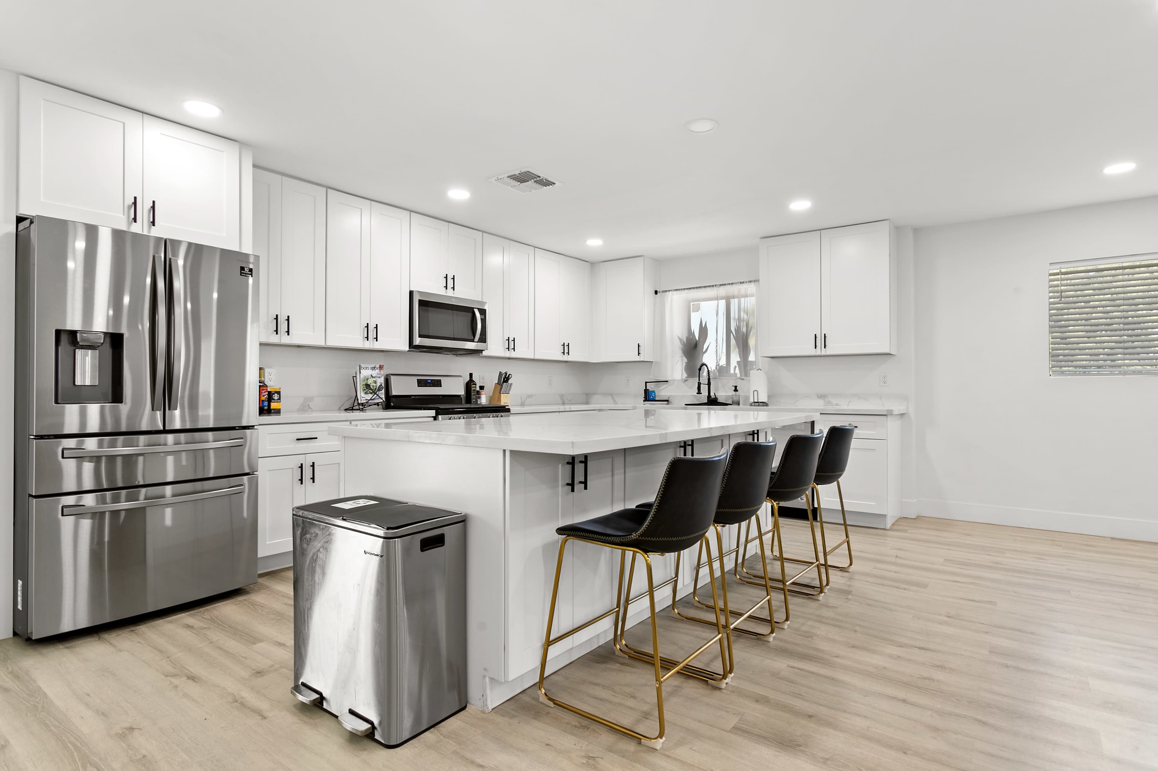 The image shows a modern kitchen with white cabinetry, stainless steel appliances, and a central island with four bar stools.