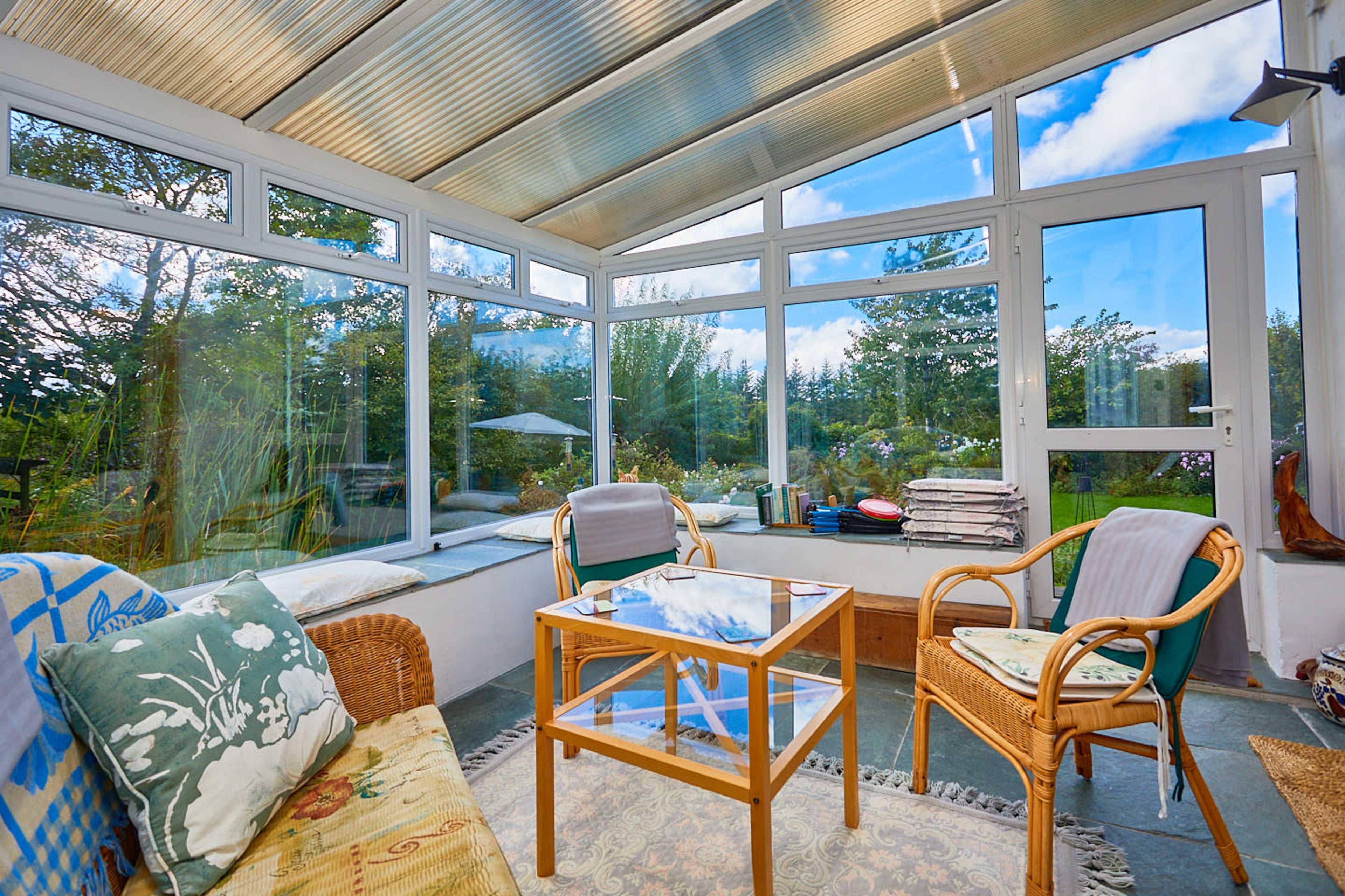 A sunlit conservatory with large glass windows, wicker furniture, and a coffee table overlooking a garden.