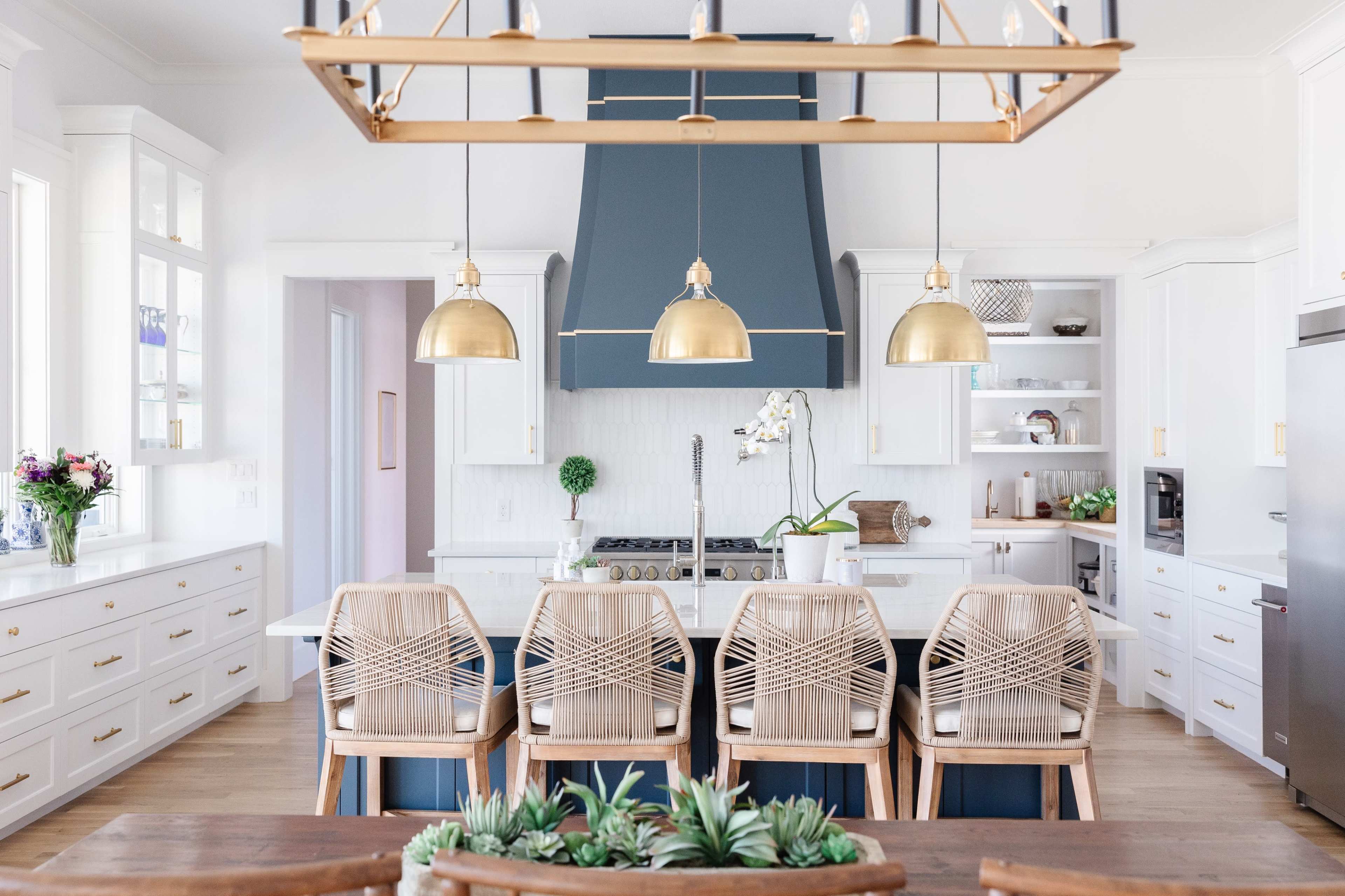 A modern kitchen with a large island, gold pendant lights, and wooden chairs arranged around a dining table.