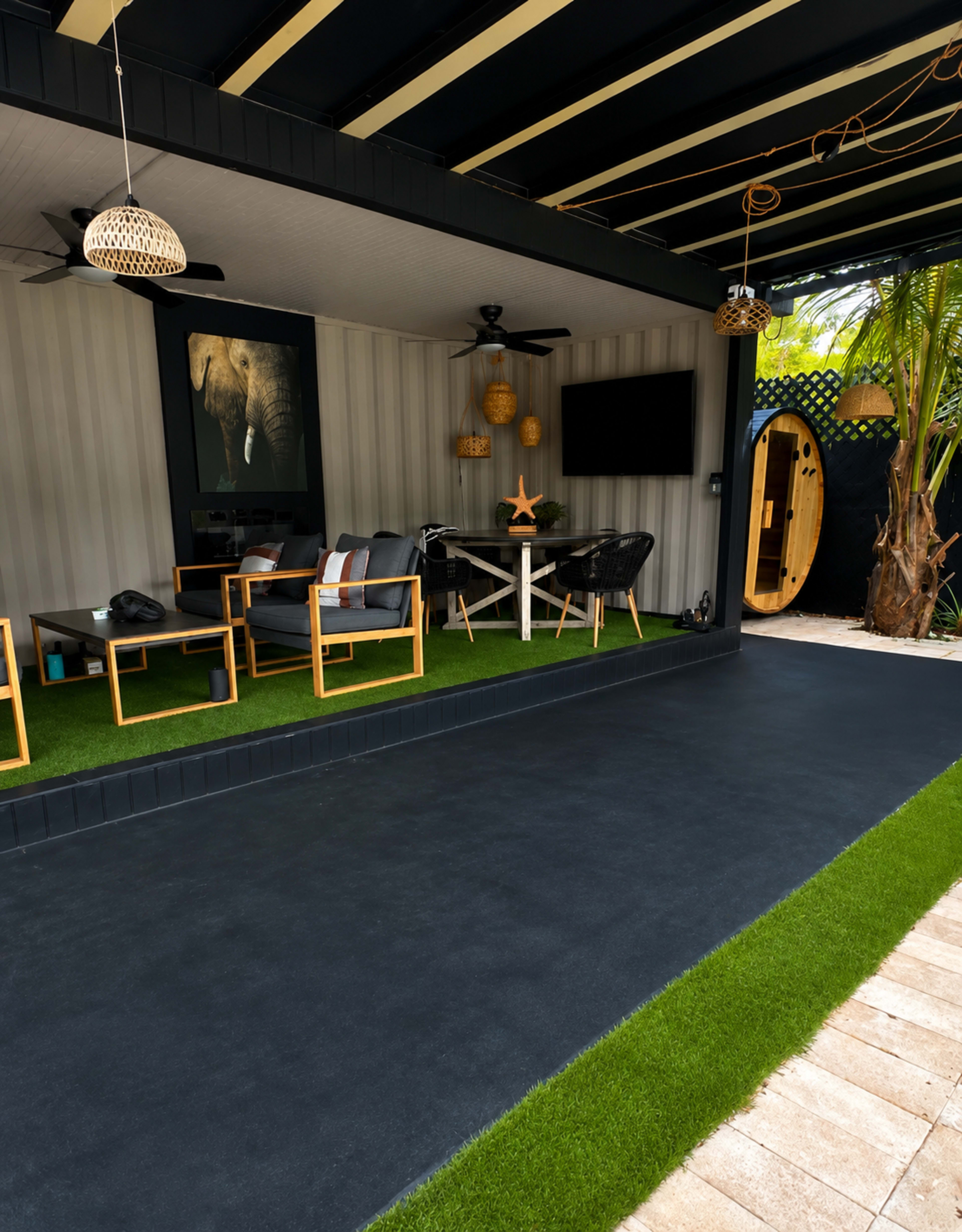 The image shows a modern outdoor lounge area with dark flooring, comfortable seating, and decorative elements including an elephant painting and tropical plants.