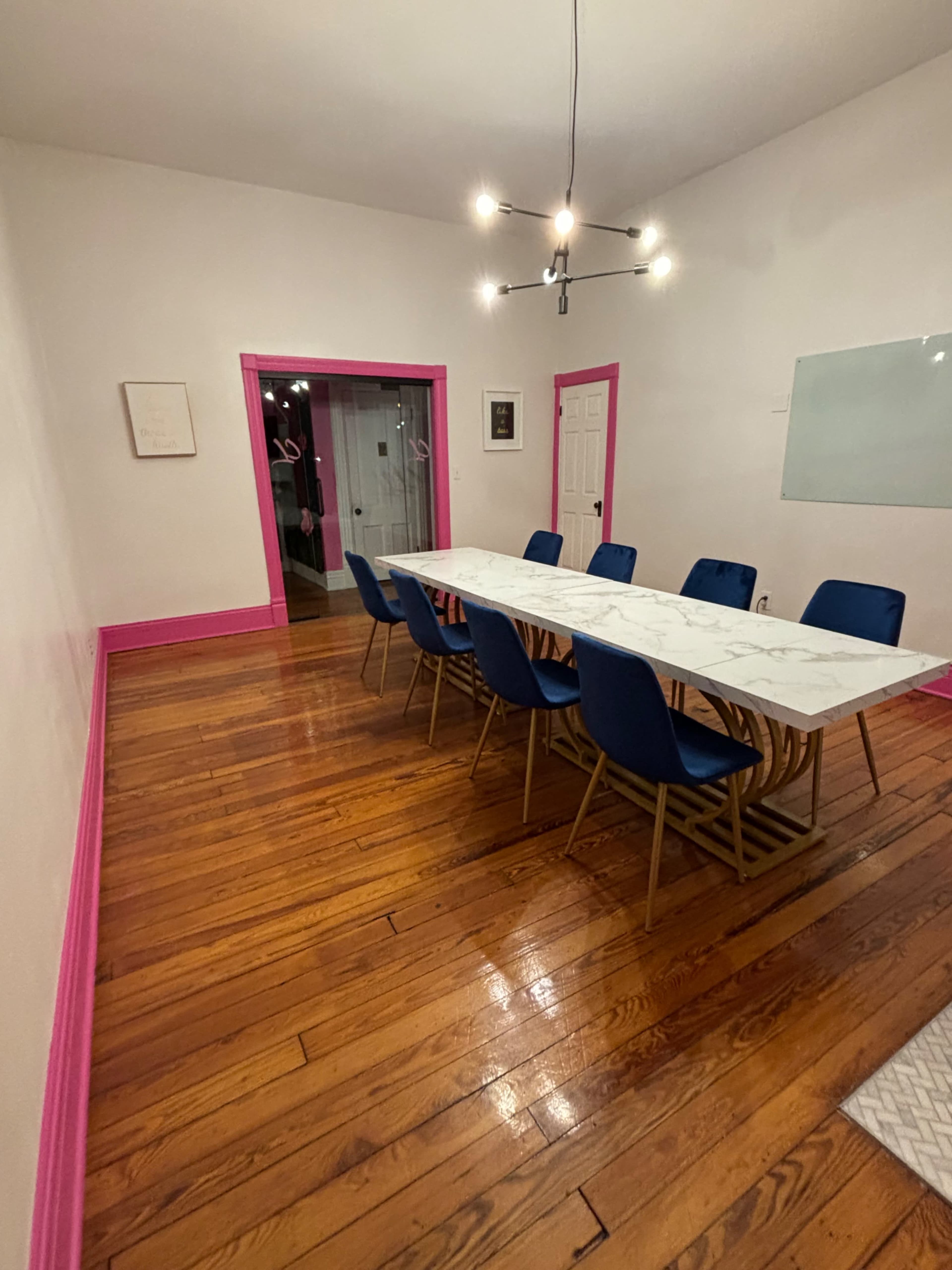A dining area with a long marble table surrounded by blue chairs, featuring pink trim along the walls and a wooden floor.
