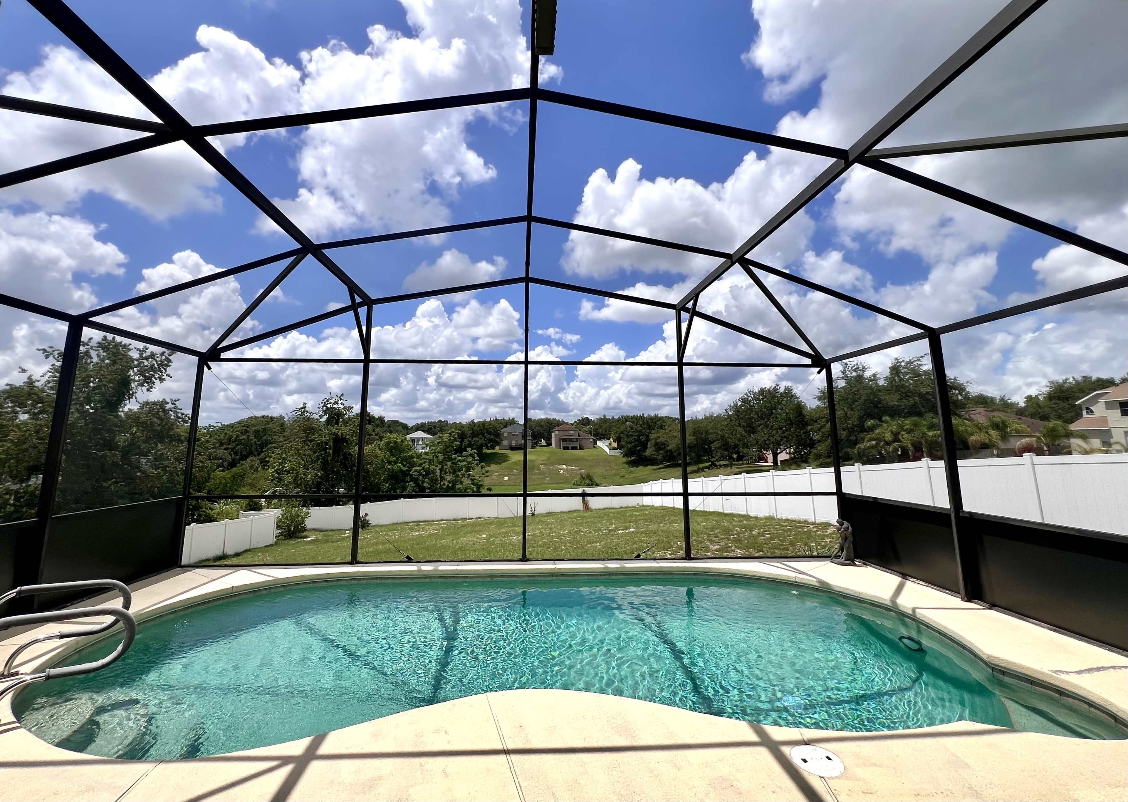 The image shows a screened-in pool area with clear water, surrounded by greenery and a blue sky filled with fluffy clouds.