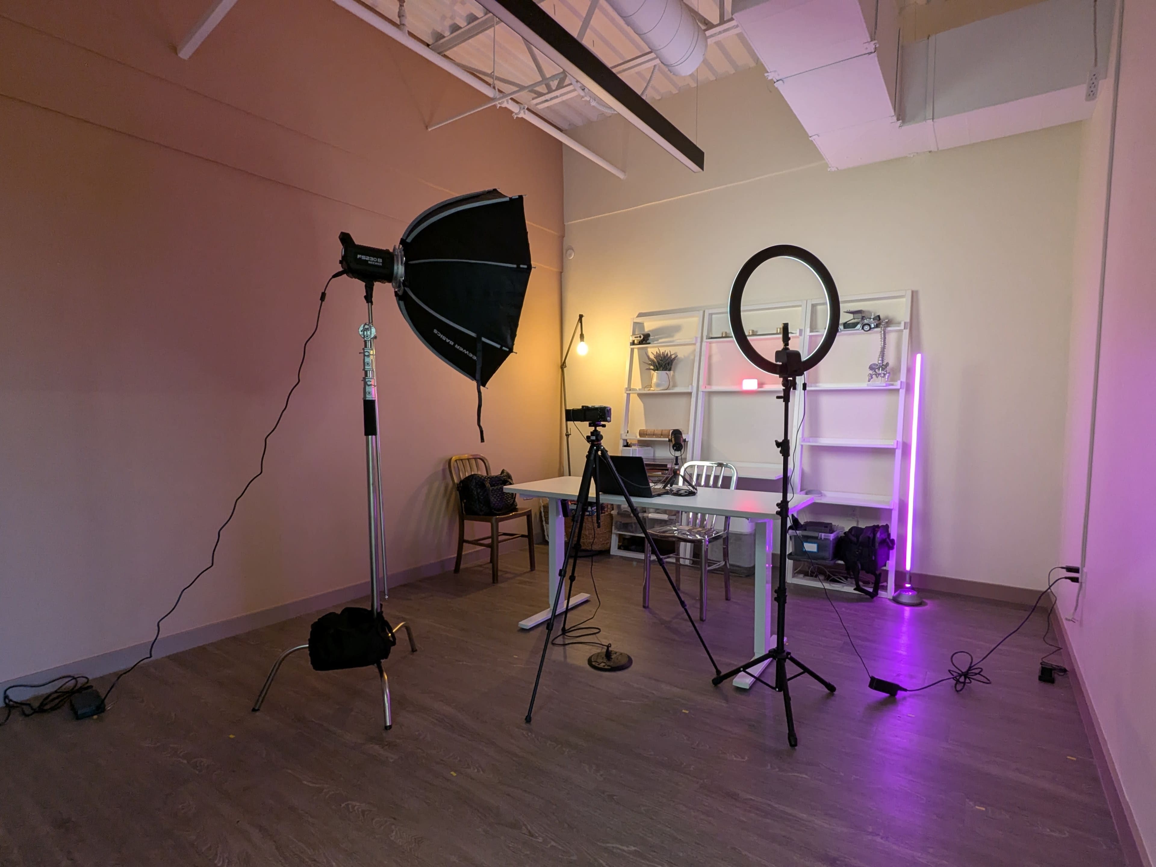 The image shows a minimalistic room set up for filming, featuring a table, various lighting equipment, and a shelf in the background.
