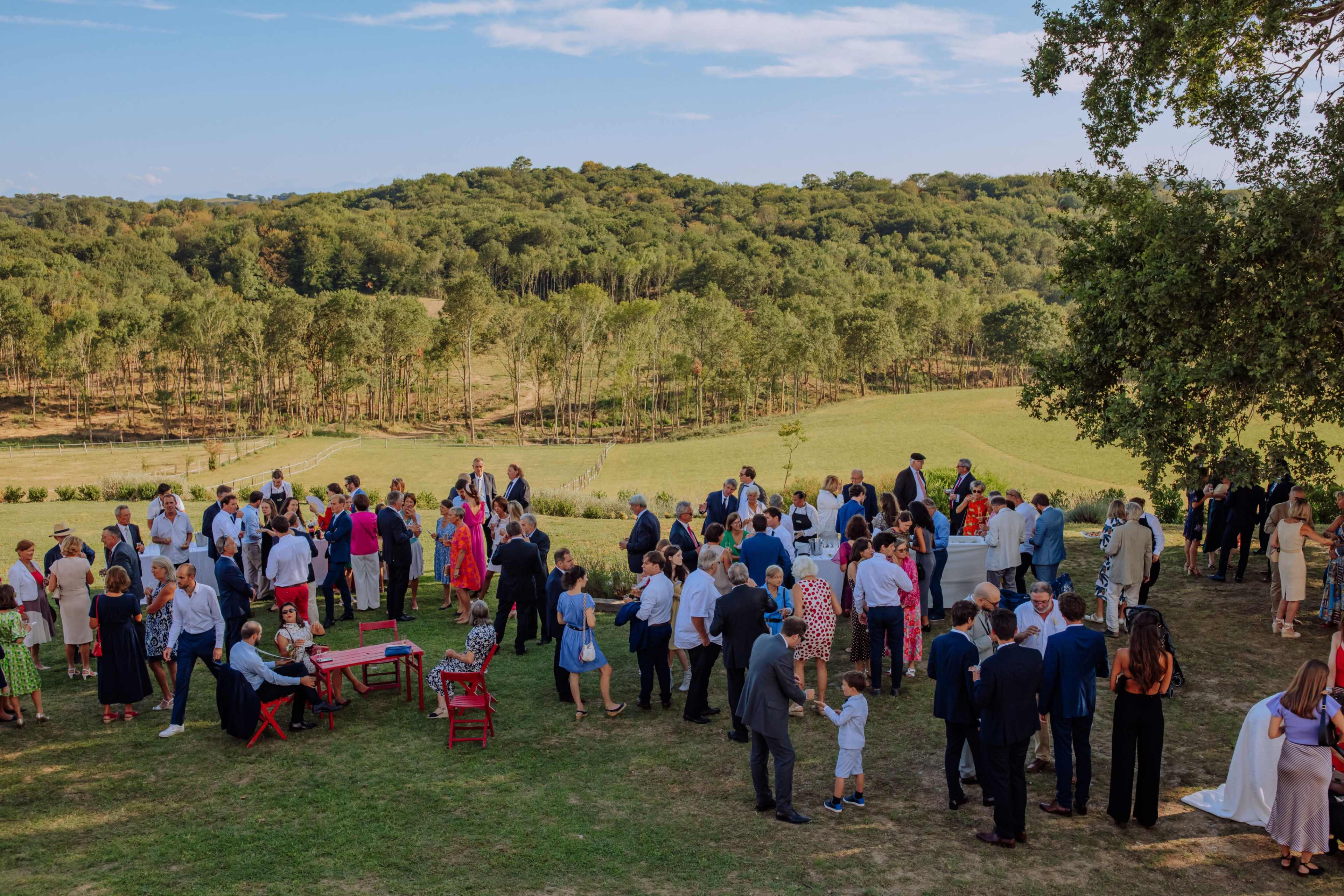 A large group of people dressed in formal attire gathers for an outdoor event on a hillside overlooking a scenic landscape.