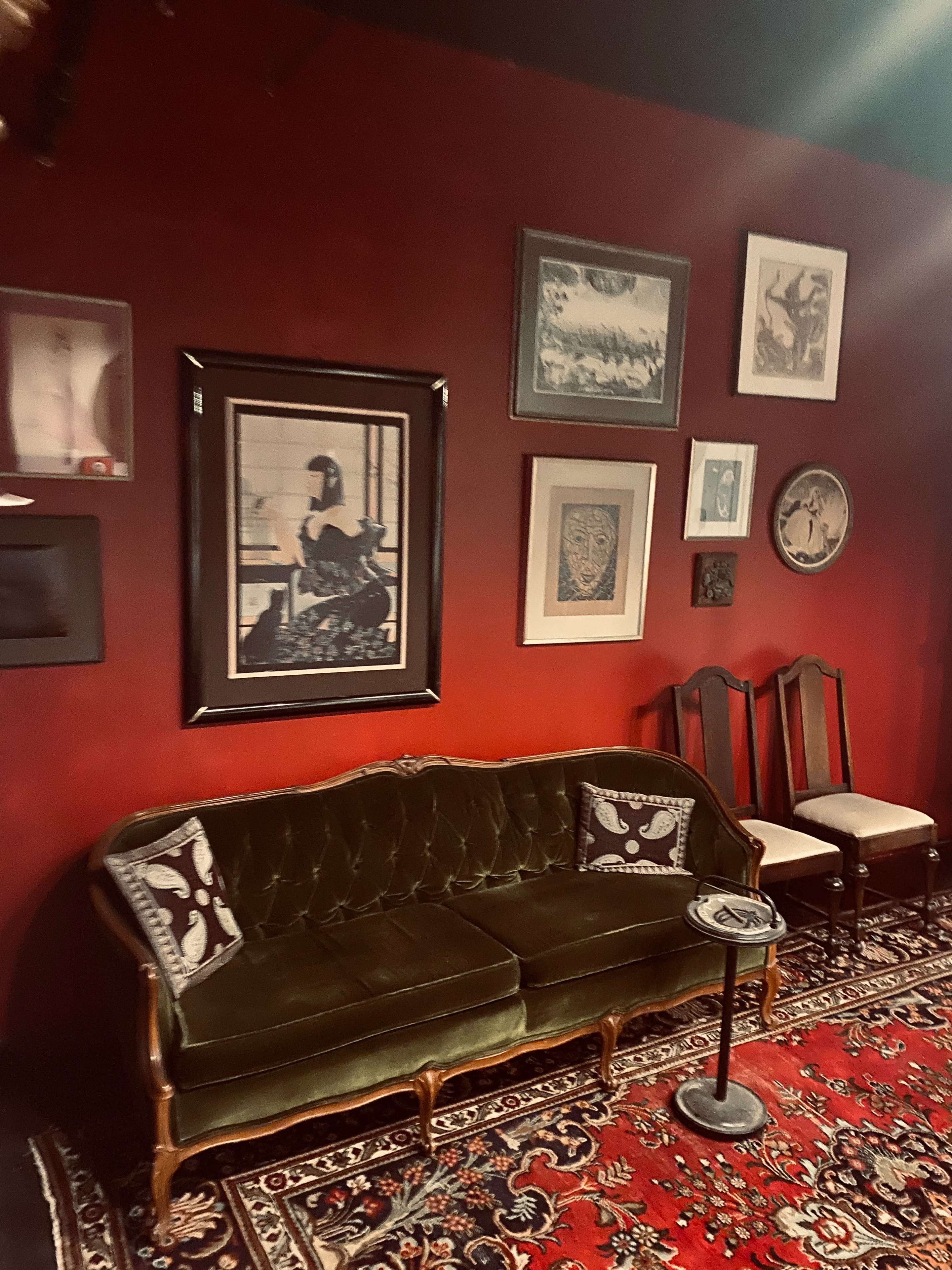 The image shows a vintage green sofa, flanked by two wooden chairs, against a red wall adorned with various framed artworks.