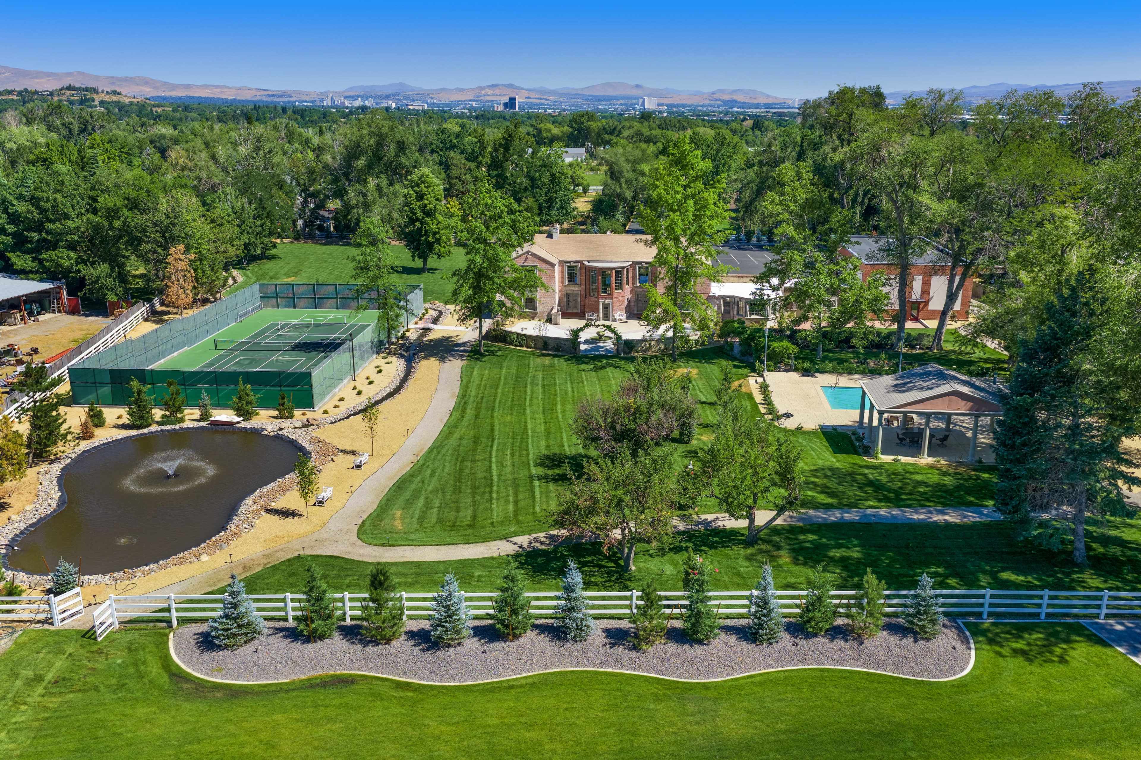 The image shows a large estate featuring a well-maintained lawn, a pond, tennis courts, a swimming pool, and a gazebo, all surrounded by trees and mountains in the background.