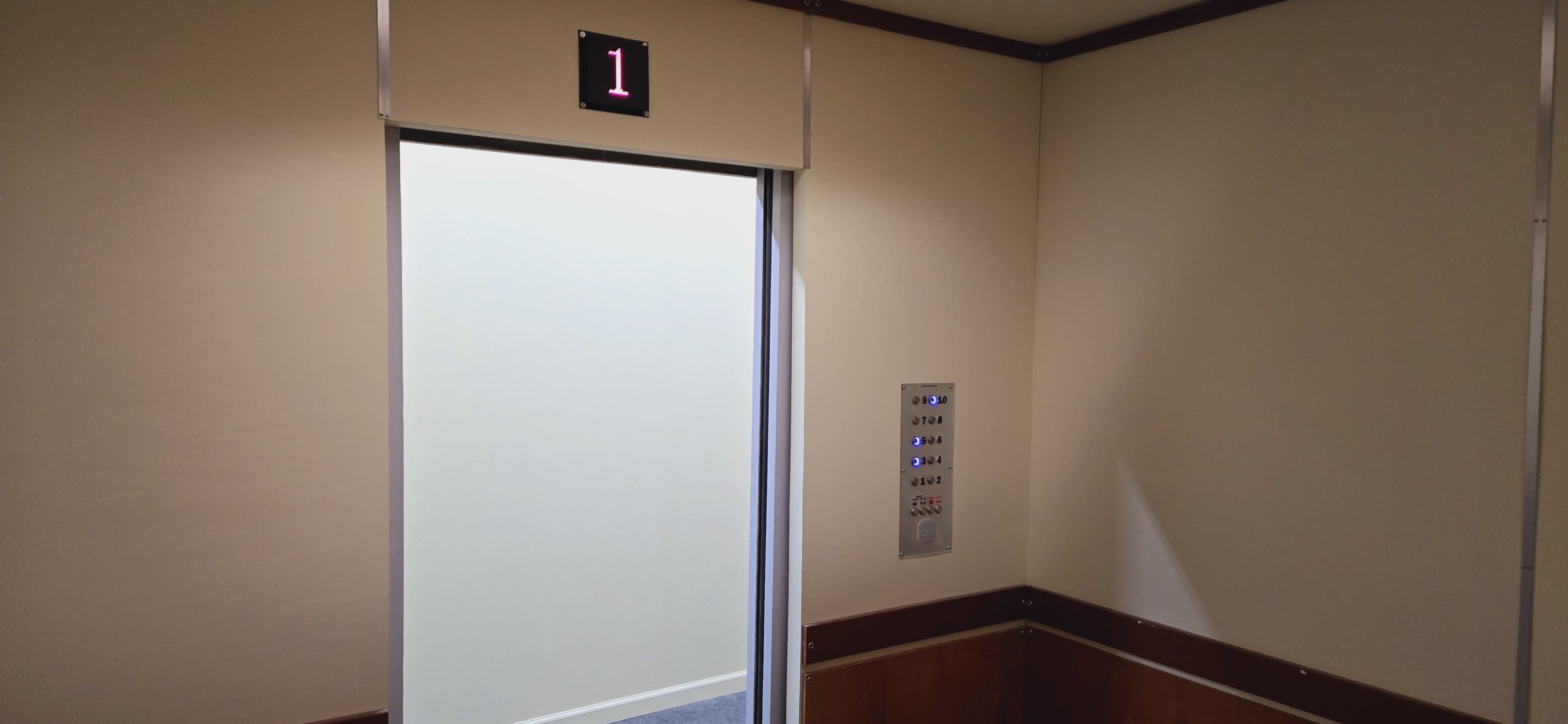 An open elevator door reveals a blank, light-colored wall with an elevator control panel beside it.