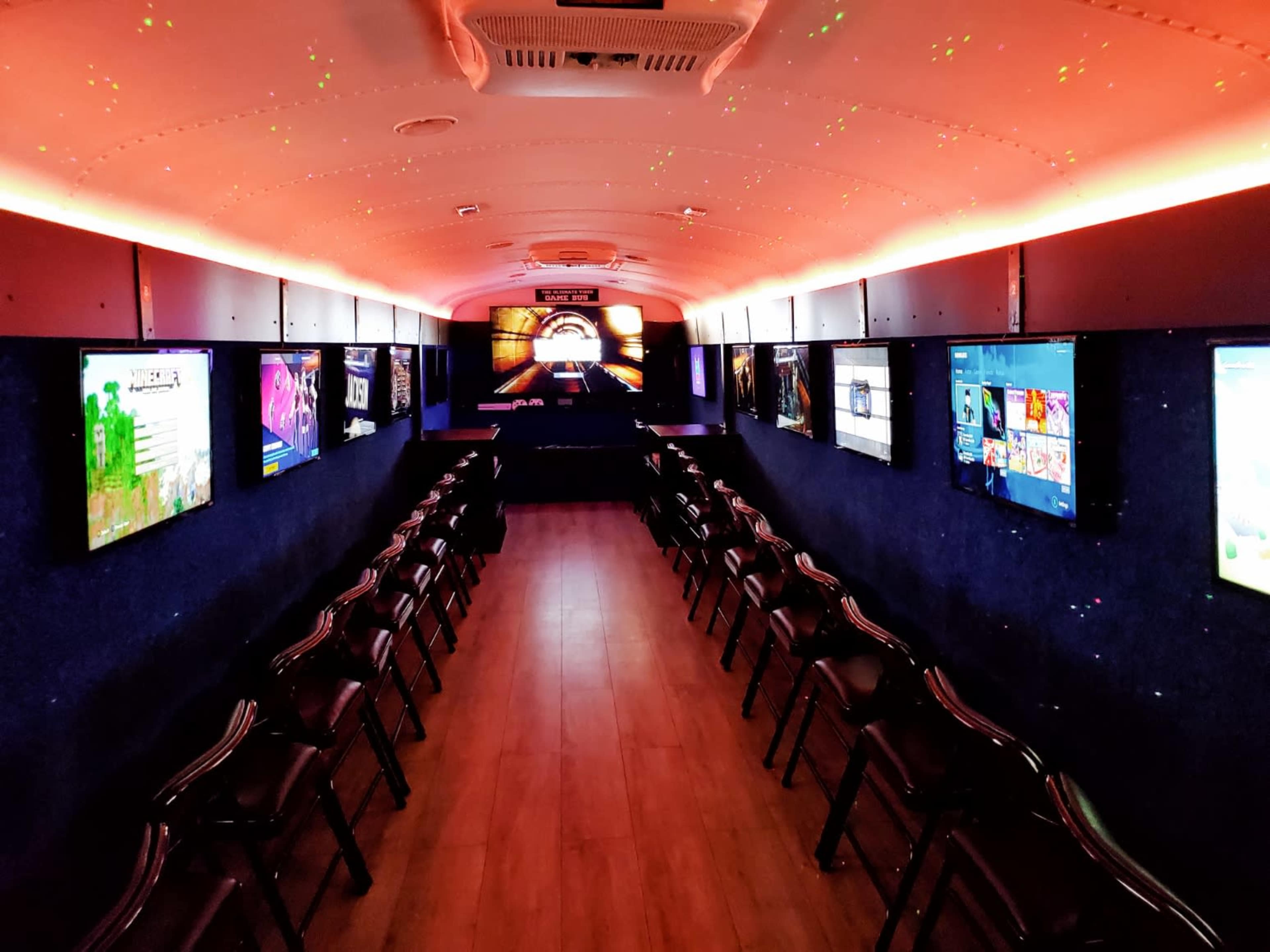 The interior of a converted bus features rows of chairs and multiple screens mounted on the walls, illuminated by colorful lighting.