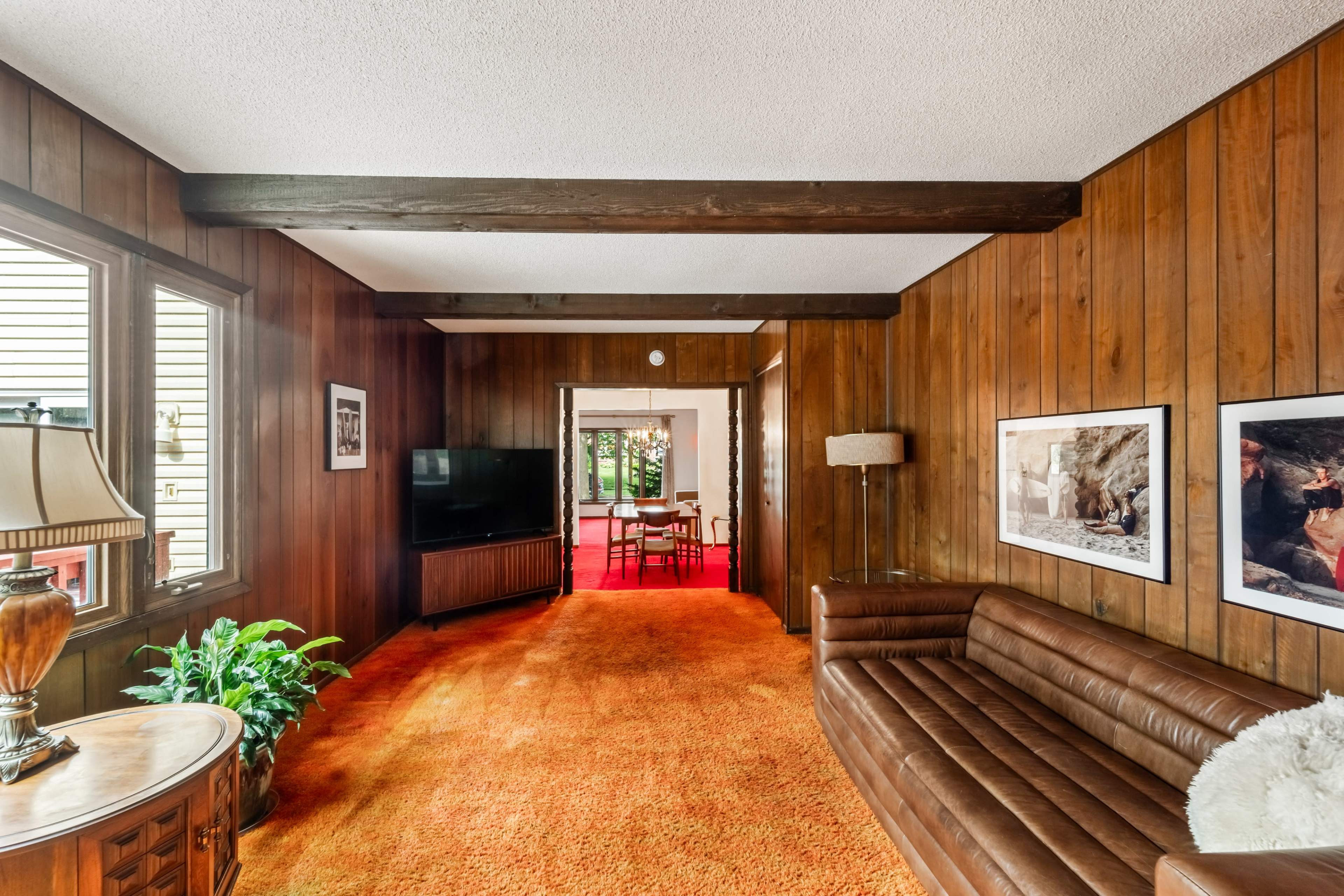 A living room with wood-paneled walls, a brown leather sofa, a standing lamp, and a television, leading to a dining area visible through an open doorway.