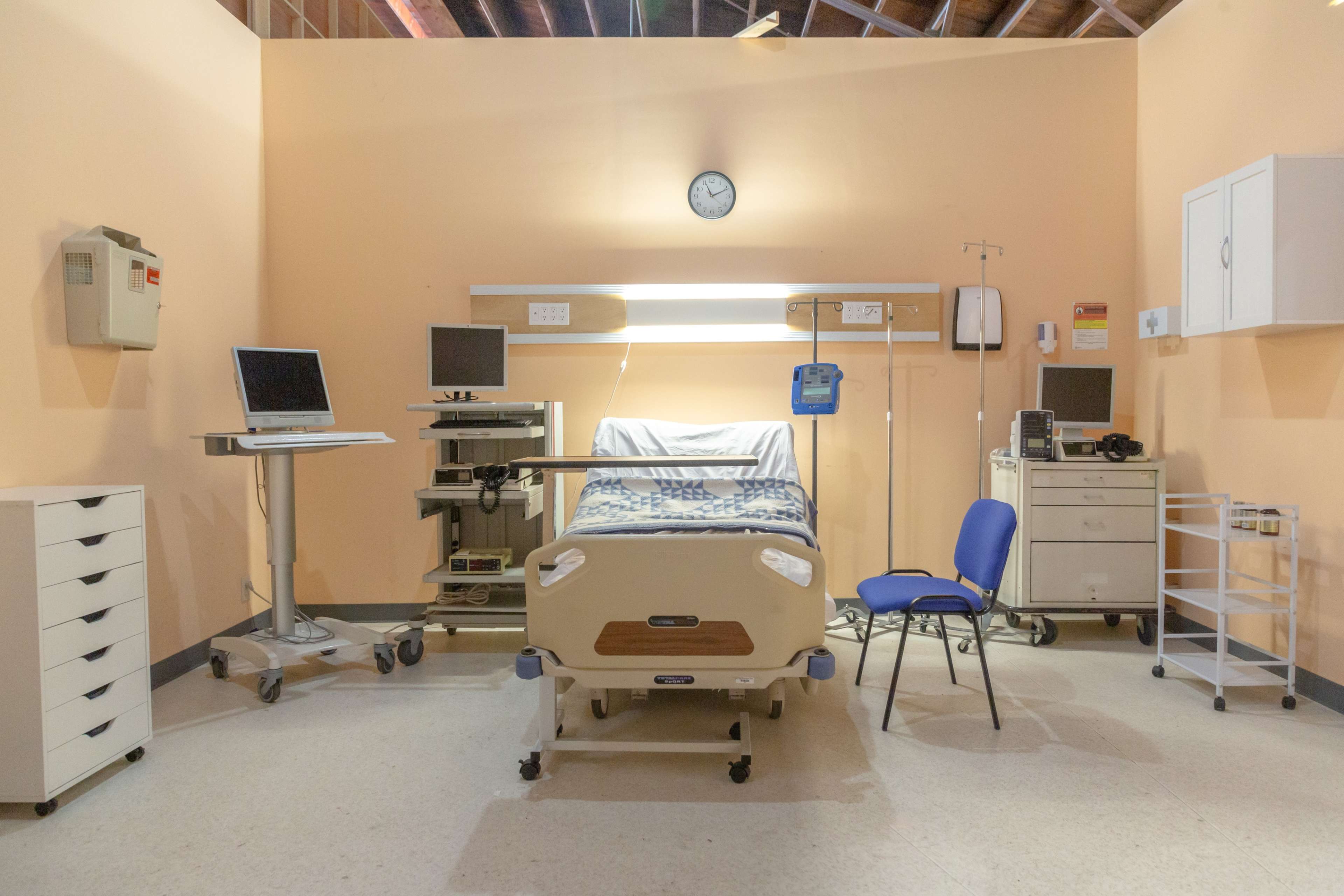 A hospital room featuring an unoccupied bed, medical equipment, a computer on a stand, and a single blue chair.