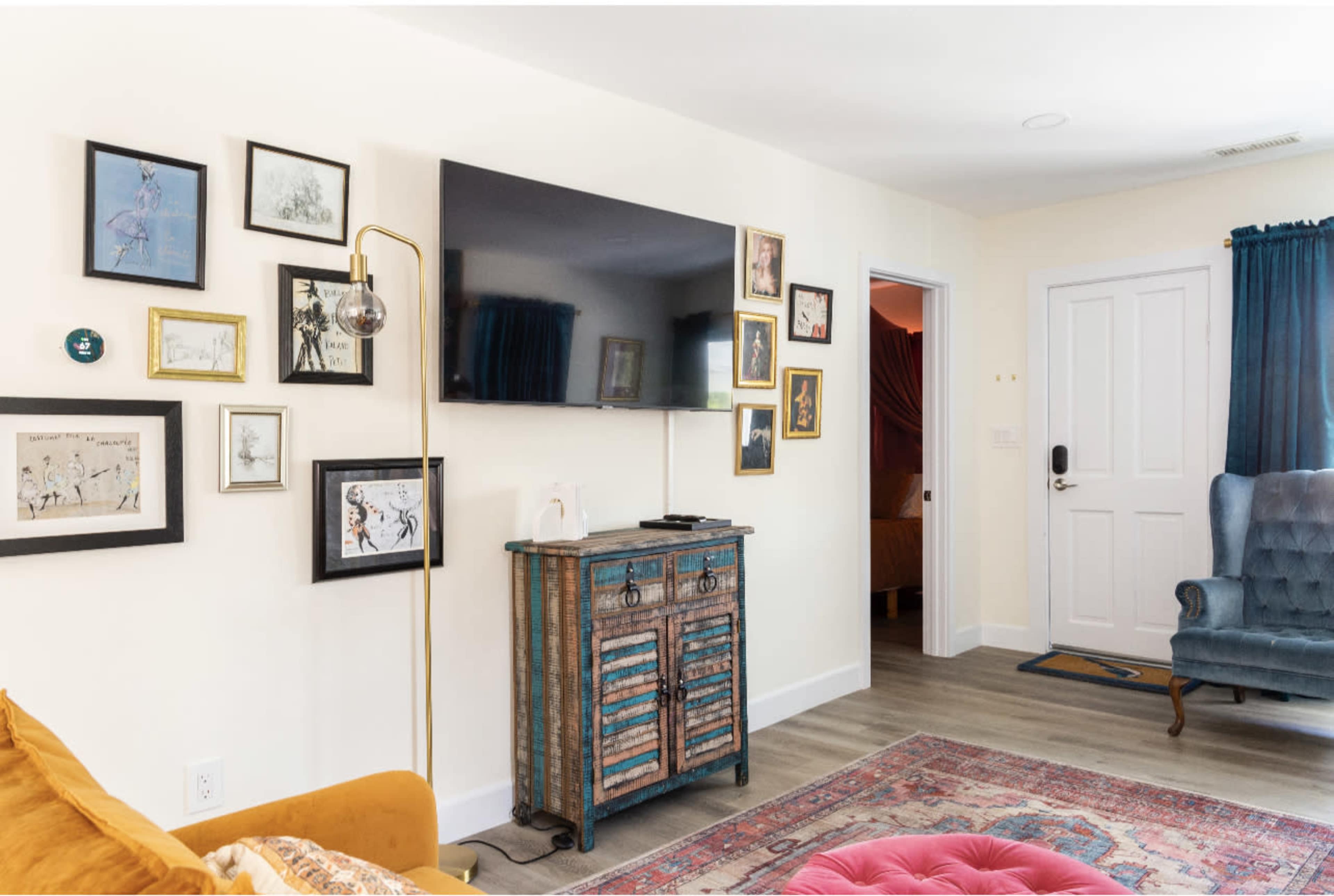 A living room features a television mounted on a wall, framed artwork arranged above a vintage cabinet, and a blue armchair beside a doorway.