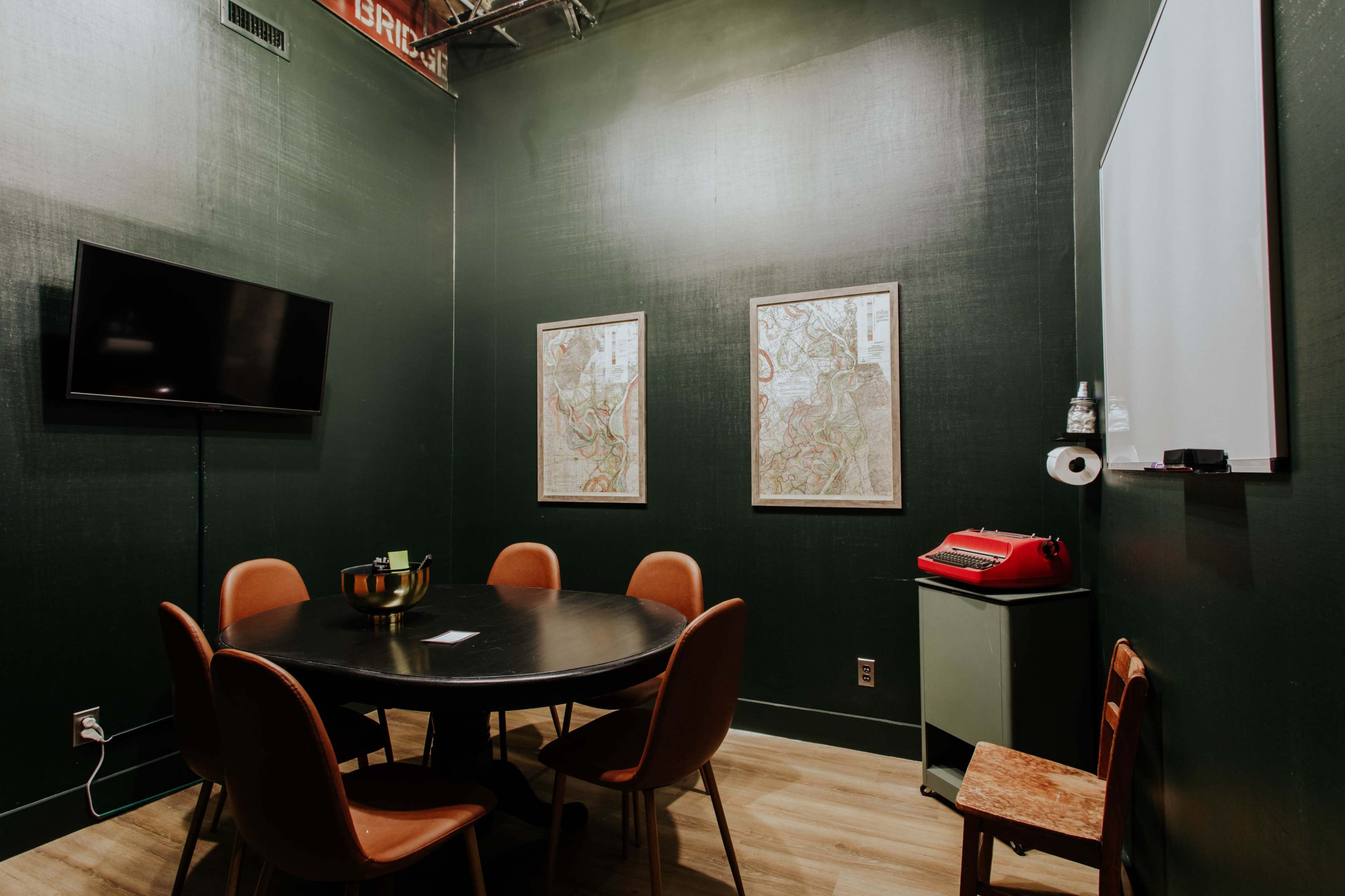 A small, dark green meeting room with a round table, four chairs, a television on the wall, and two framed maps.