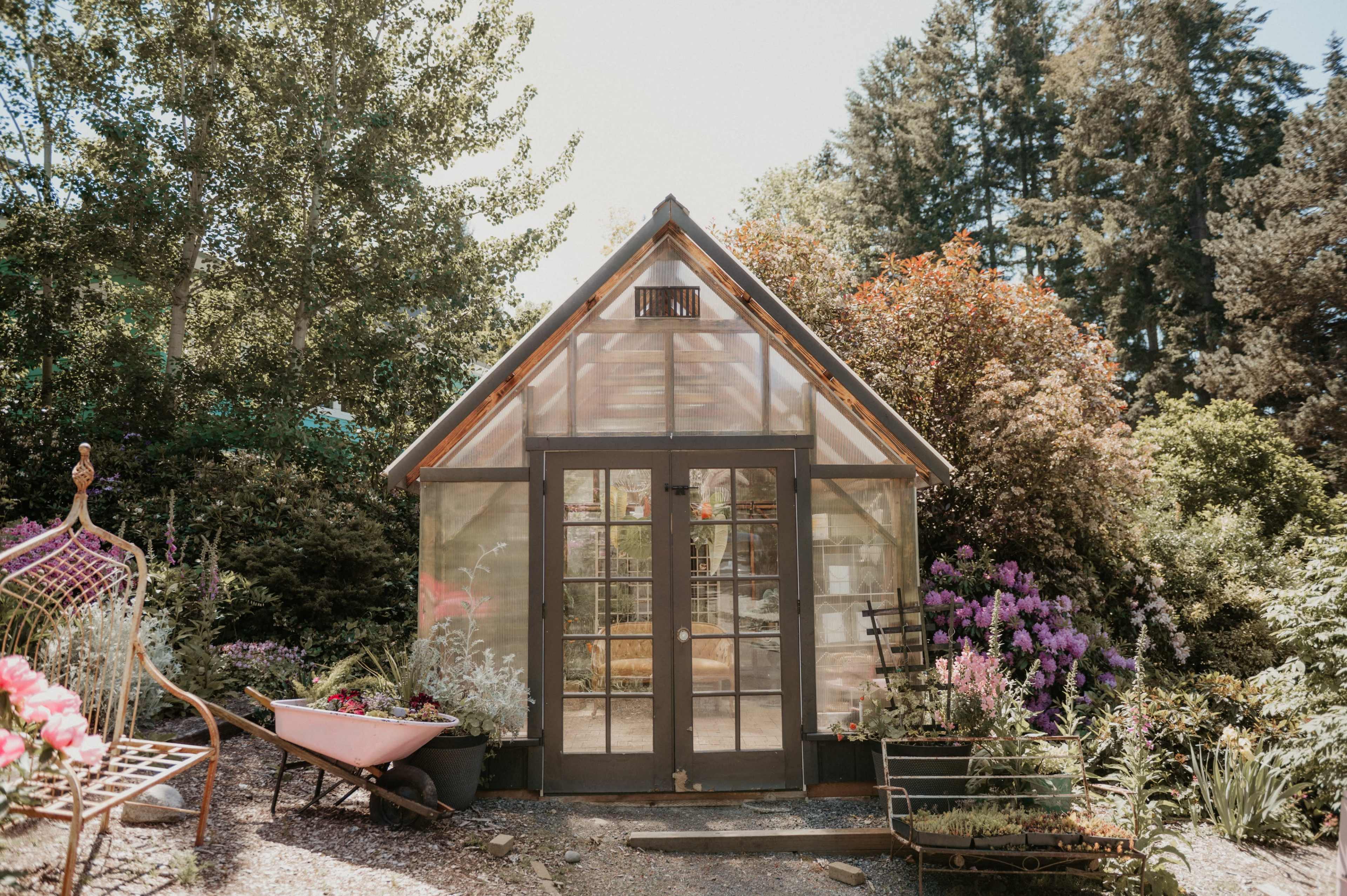 A greenhouse with glass walls and a peaked roof is surrounded by vibrant flowers and greenery.