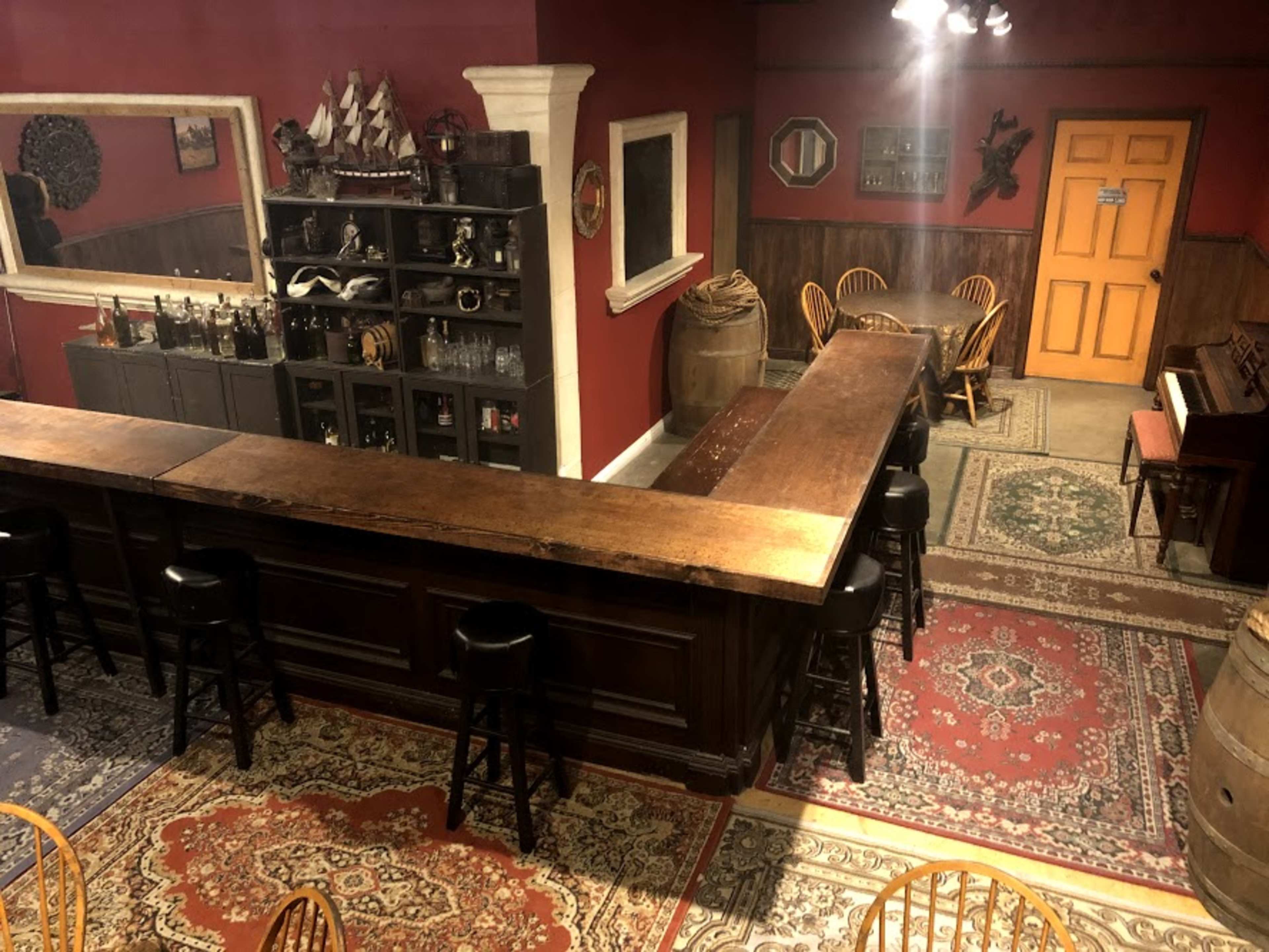 A dimly lit bar area with a wooden counter, shelves of bottles, and a piano, all set against a red wall and patterned rugs.