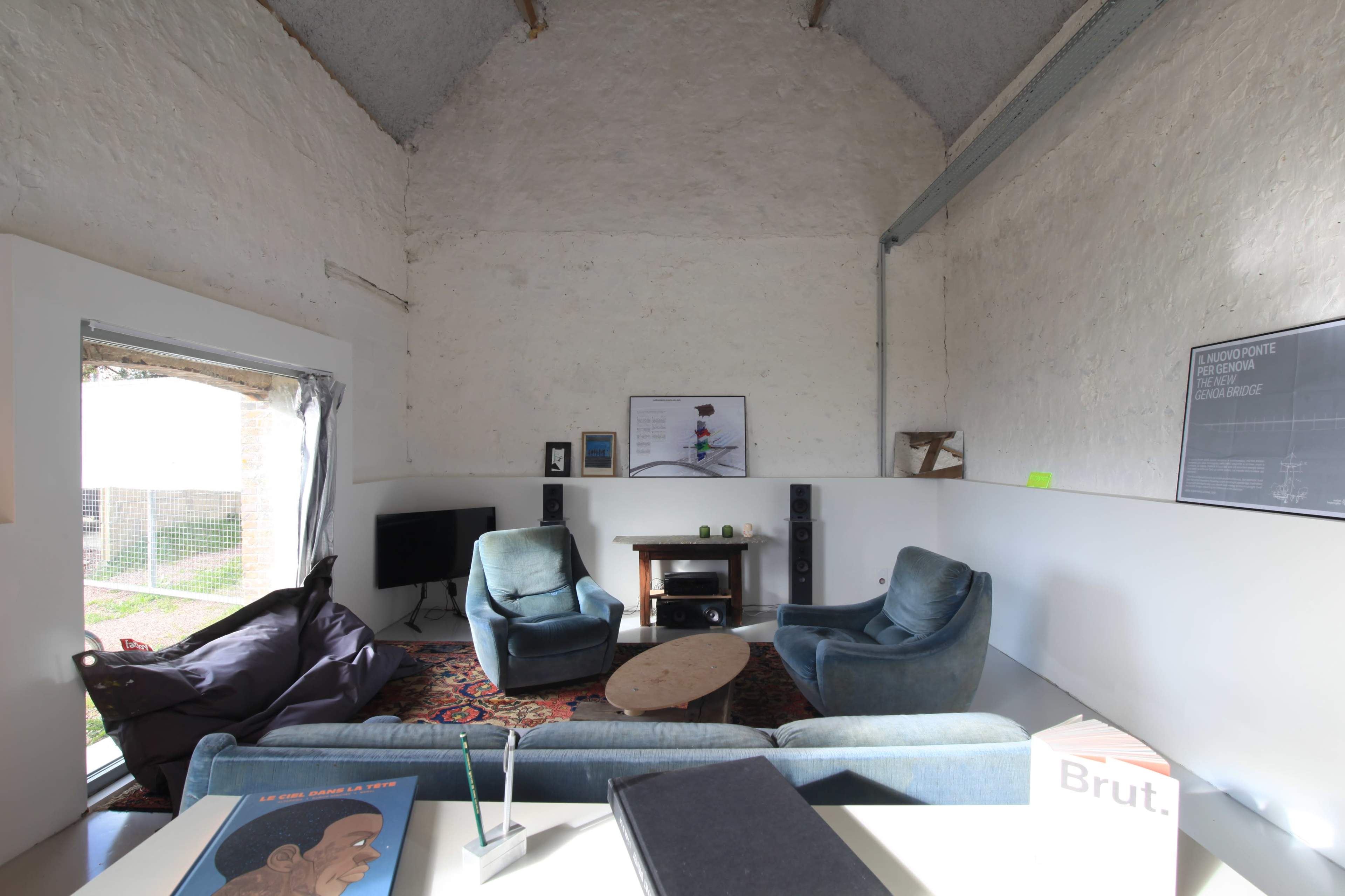 A spacious, minimalist living room with two blue armchairs, a circular wooden coffee table, a television, and a view of an outdoor area through a large open window.