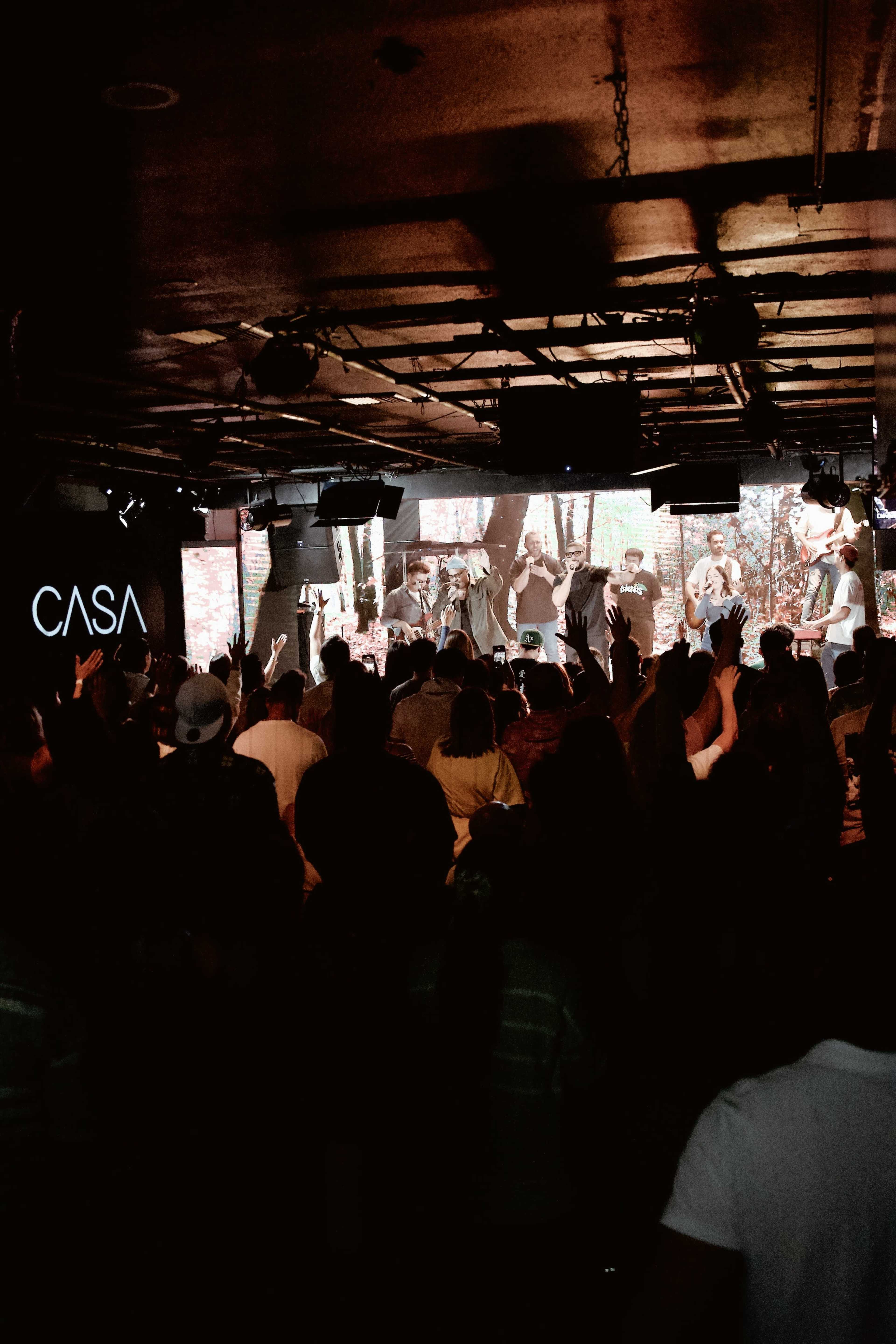 A crowd of people is gathered in a dimly lit venue with a large display screen featuring a performance on stage.