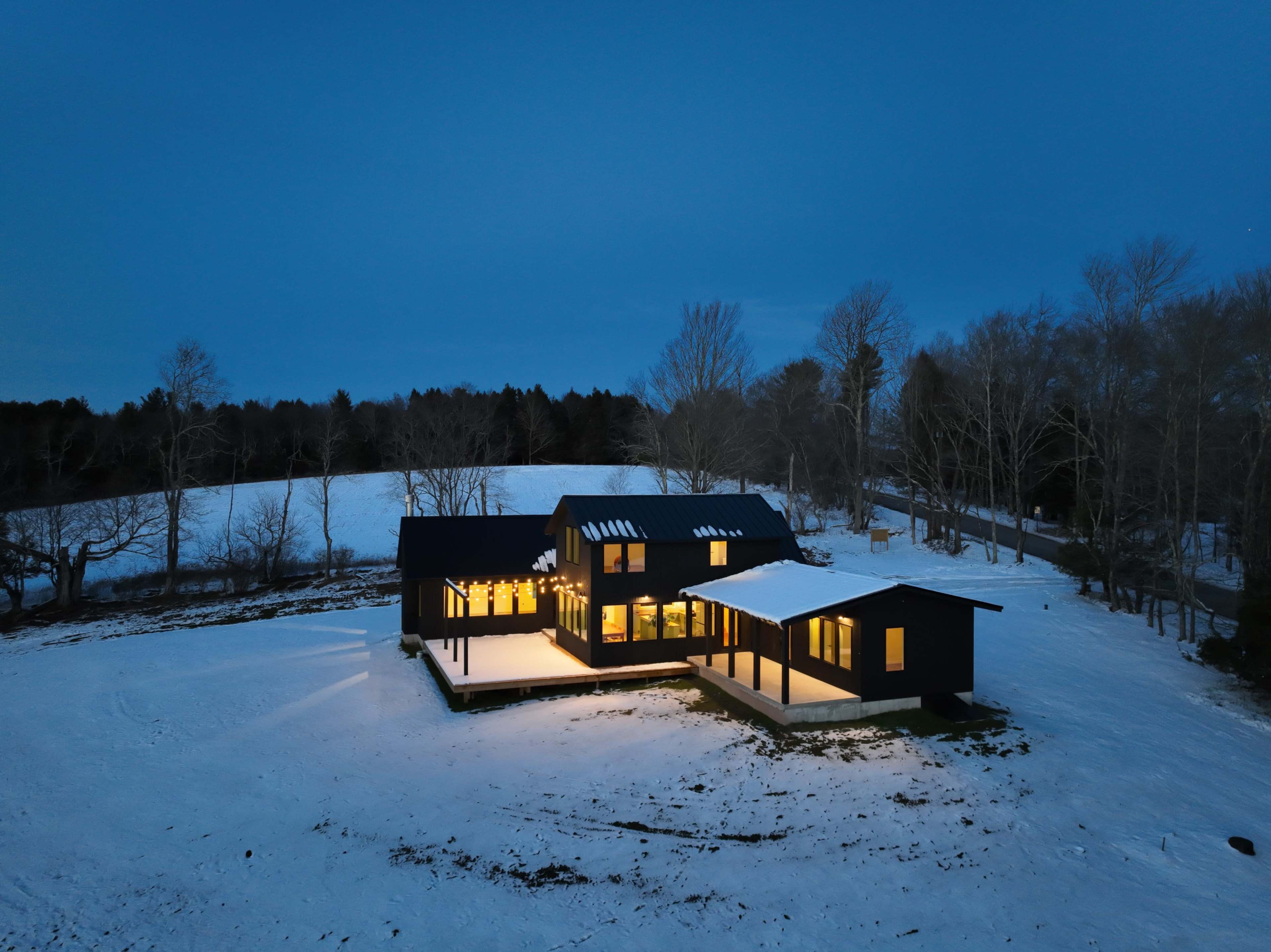 A modern black house with large windows is surrounded by snow-covered land under a twilight sky.