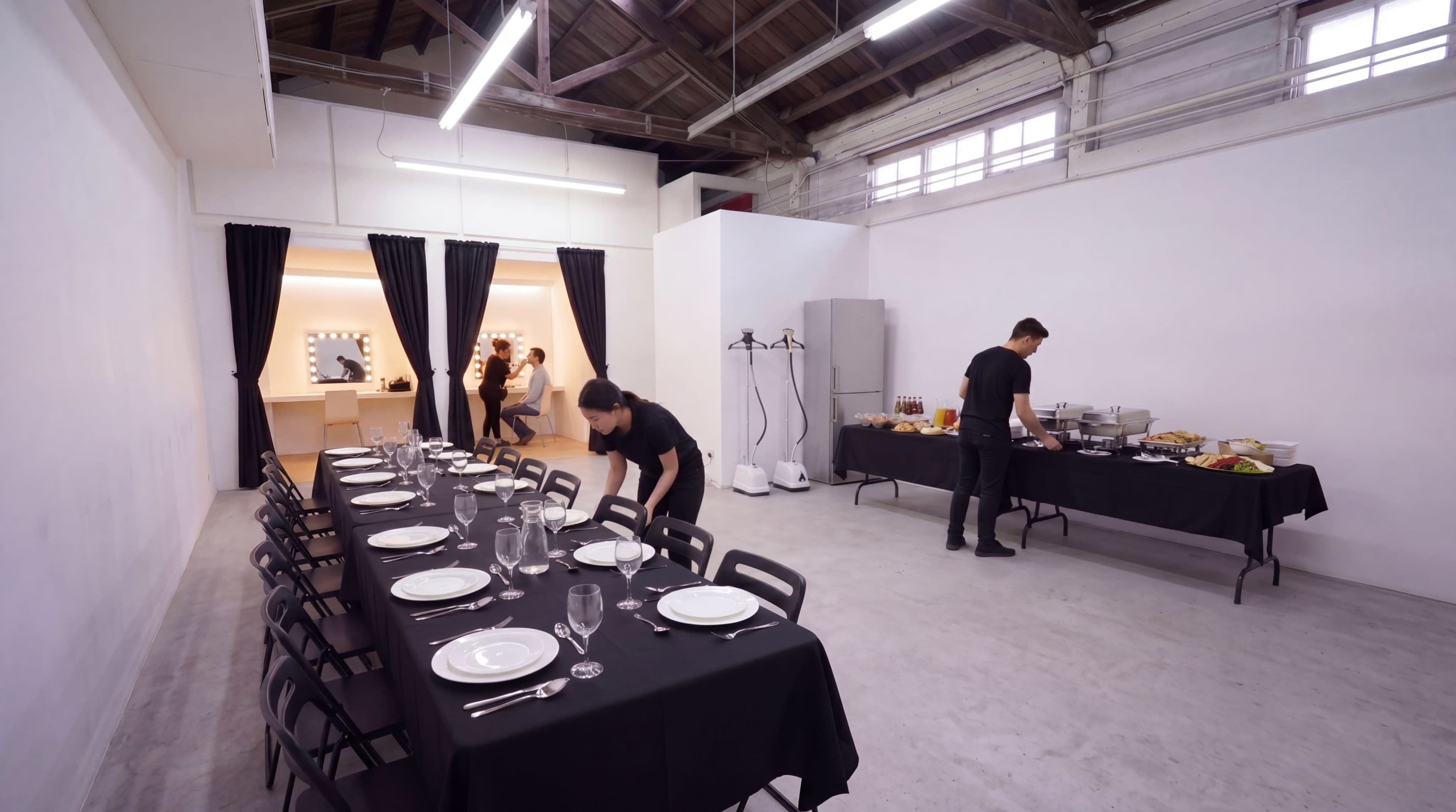 A large dining space features a long black table set with plates and glassware, while staff prepare a buffet on a separate table and attendees are visible in the background.