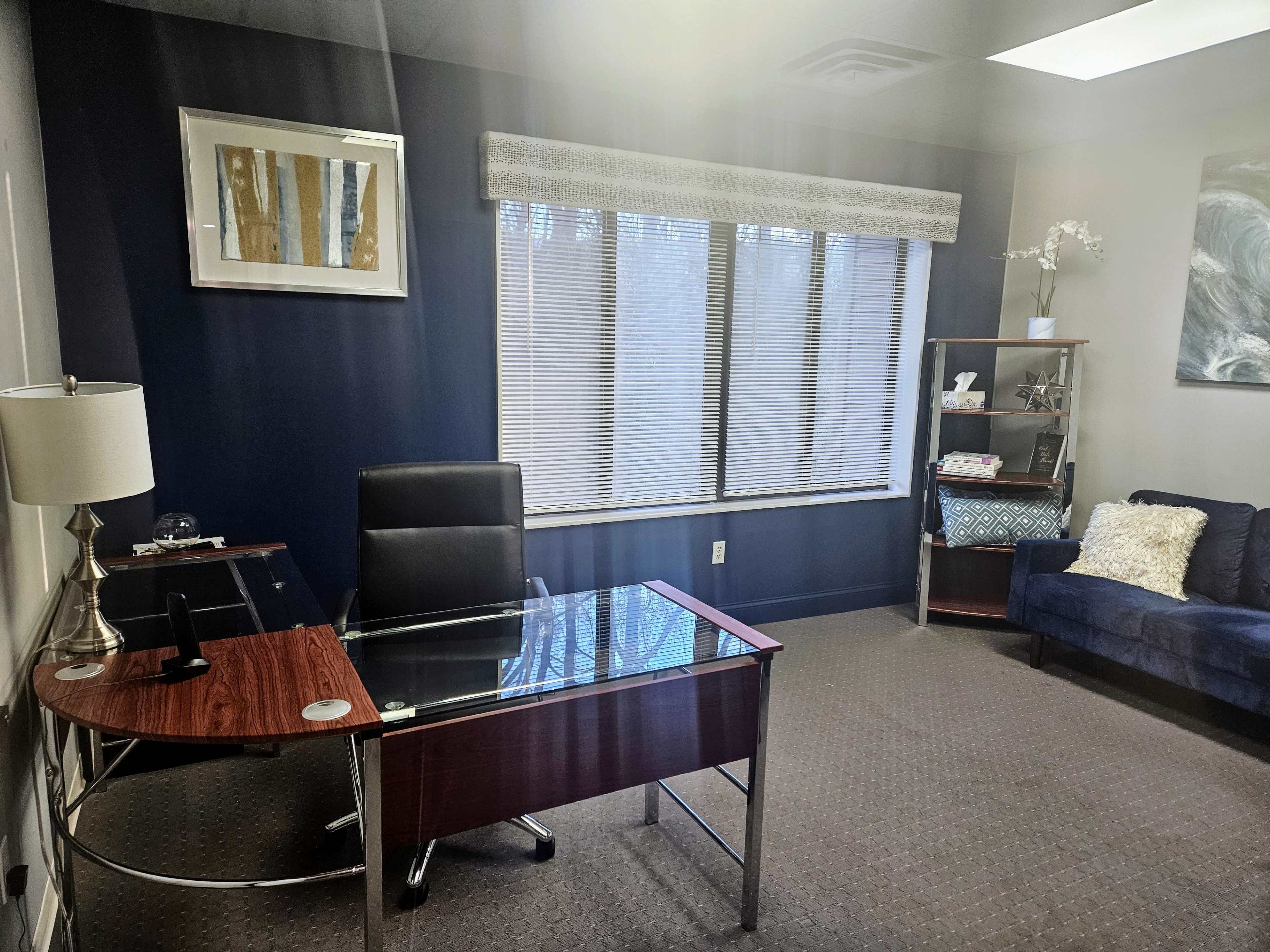 An office space featuring a glass-top desk, a black chair, a bookshelf, and a blue sofa, with large windows allowing light to enter.