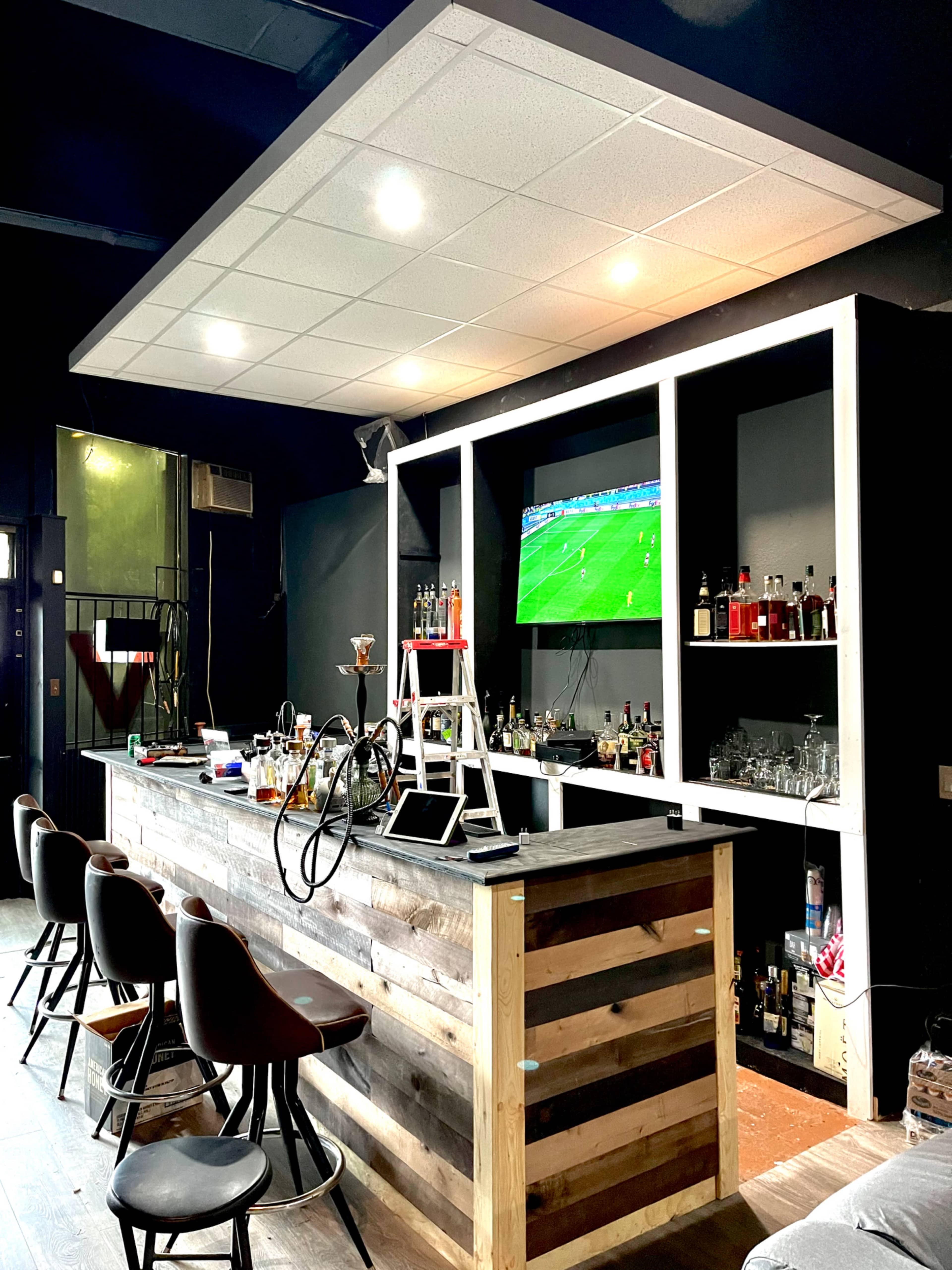 The image shows a modern bar with a wooden counter, several bar stools, and a television displaying a sports game in a dimly lit interior.