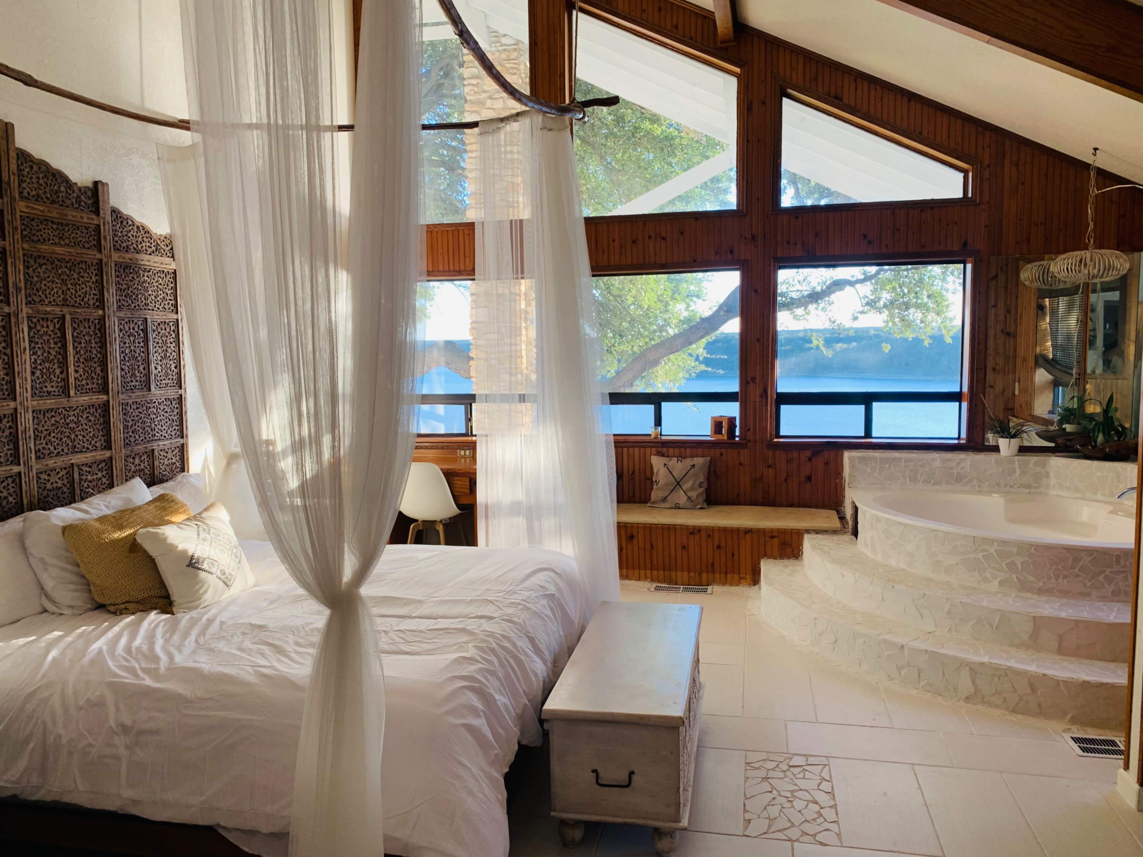 A spacious bedroom features a bed draped with sheer curtains, large windows overlooking a body of water, and a circular jacuzzi on a raised platform.