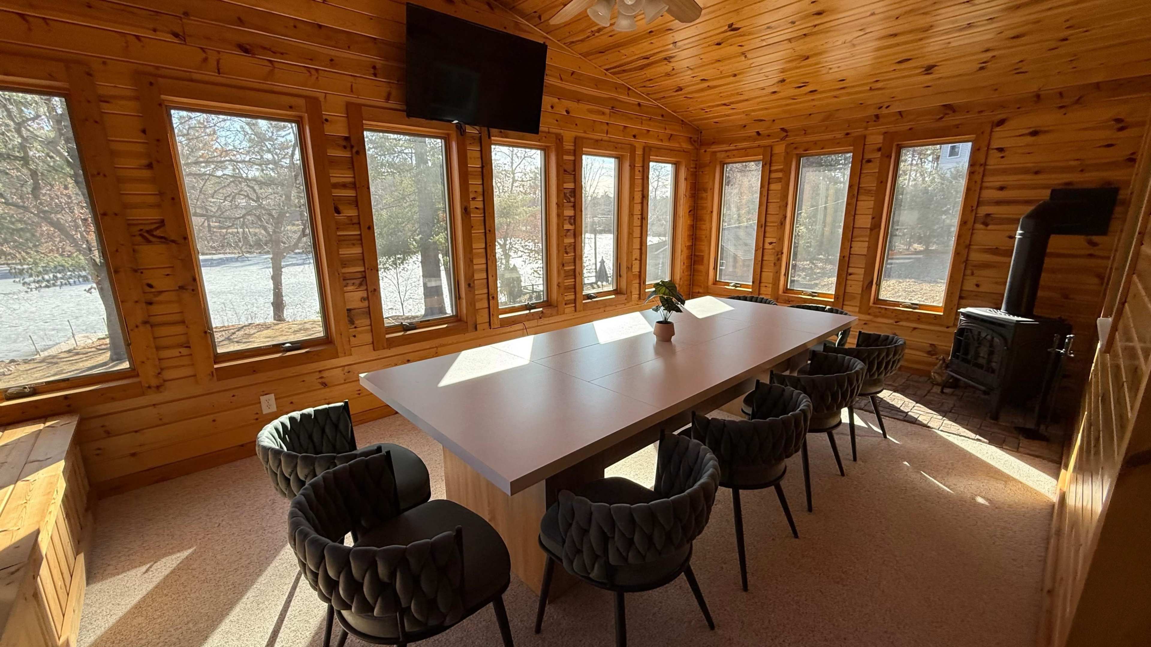 The image shows a sunlit wooden room with large windows, featuring a long table surrounded by eight black chairs and a small plant in the center.